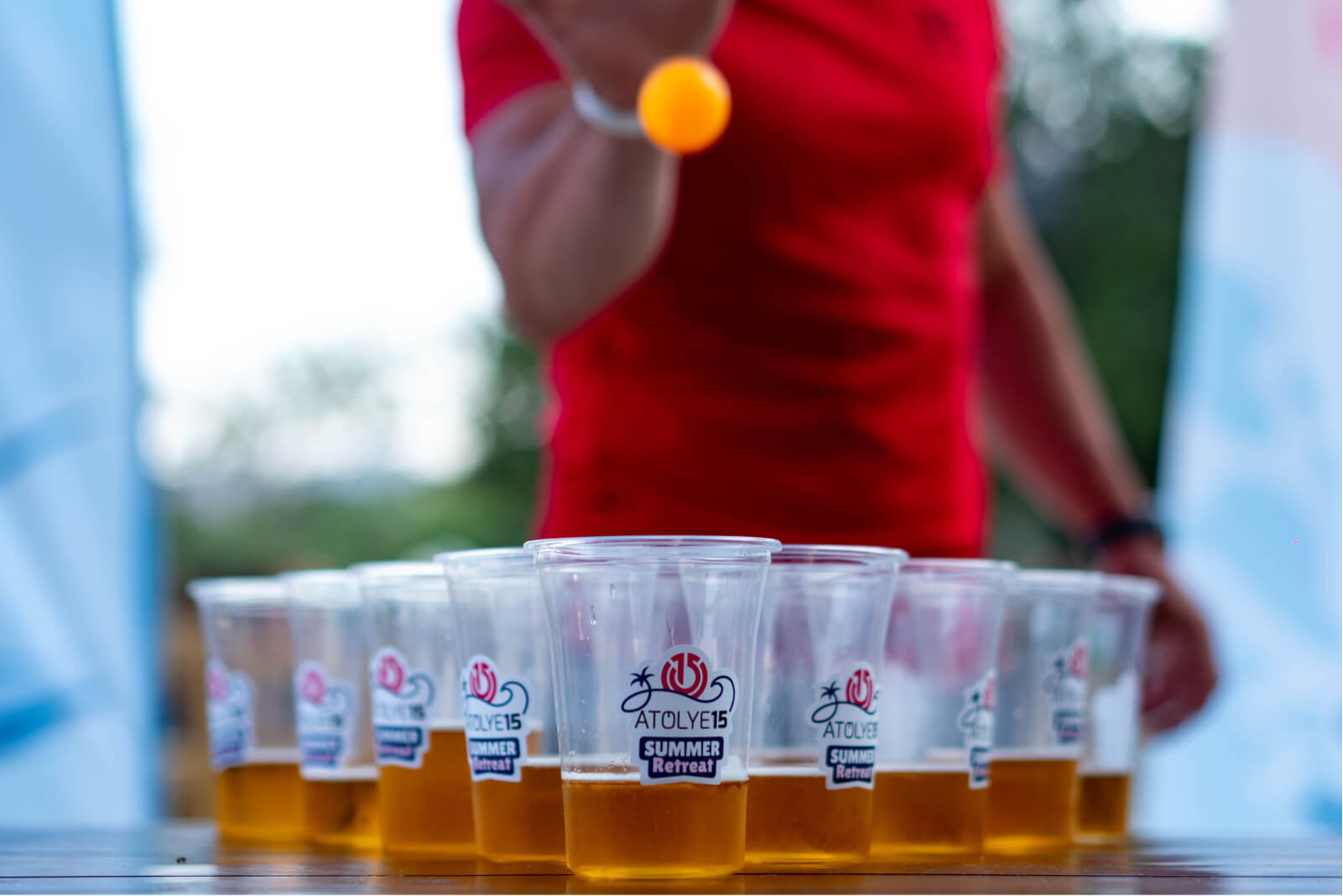 A man in a red t-shirt playing beer pong, throwing an orange ball into plastic beer cups that say “Atolye15 Summer Retreat.”