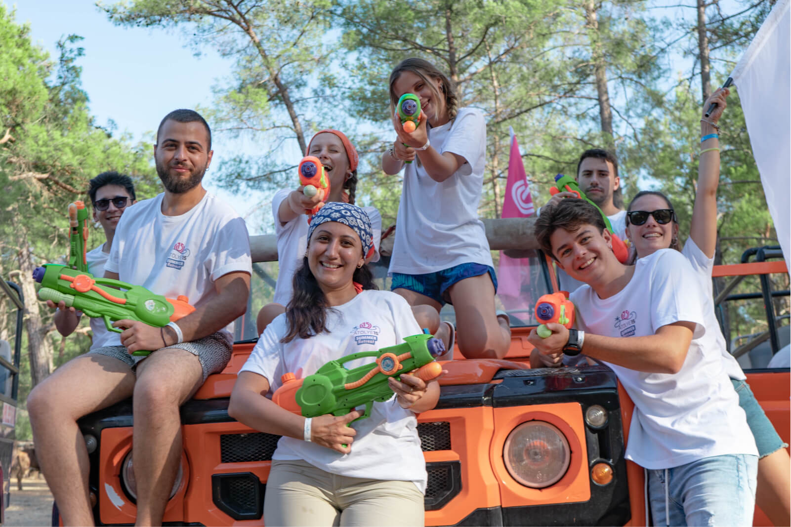 A group of people gathered around an orange jeep posing with green water guns in front of the Atolye15 logo.
