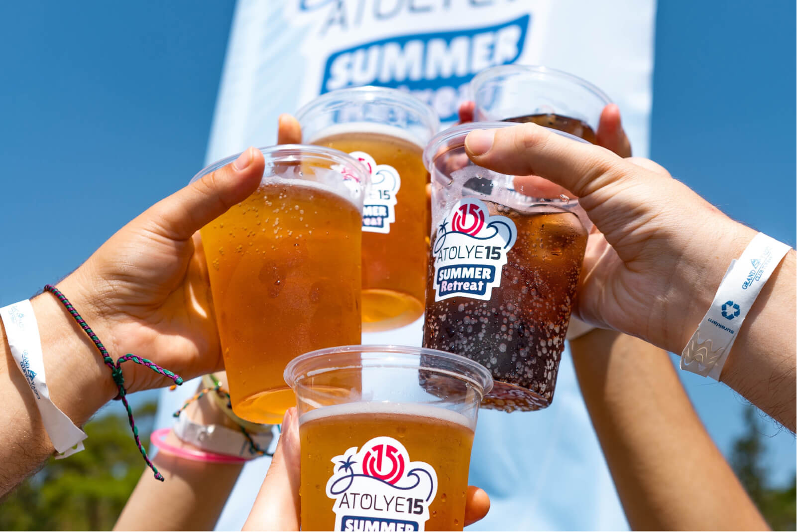 Wristbands around hands that raise plastic beer cups that show the Atolye15 logo in front of a white Summer Retreat banner.