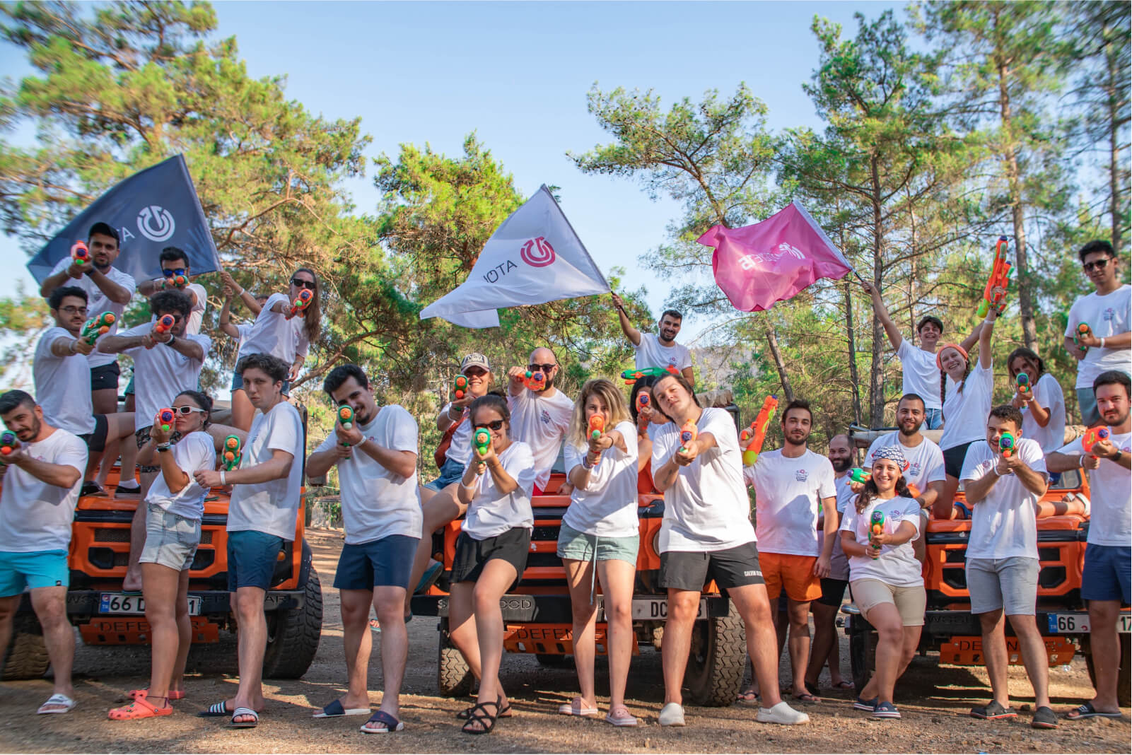 A group of people, wearing white t-shirts and summer shorts, posing with water guns, and waving Atolye15 flags.