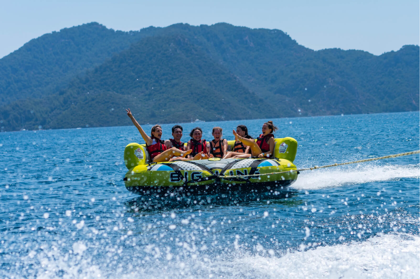 Five people on a green inflatable boat in the sea by a mountain.
