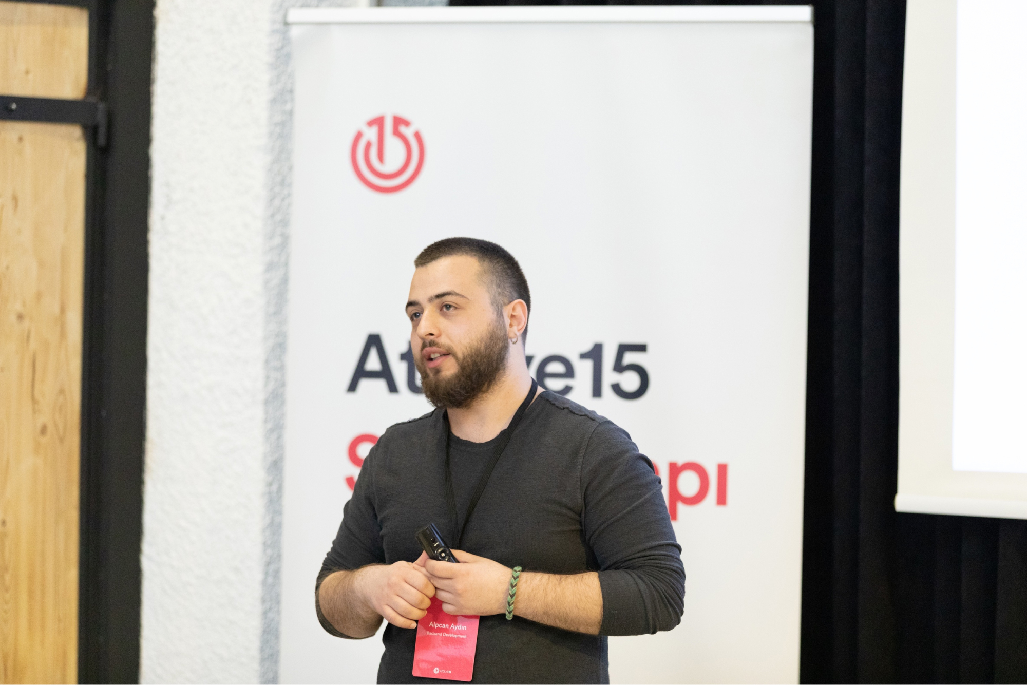 A bearded man in a dark grey sweatshirt, talking in front of the white “Staj Kampı” banner with the Atolye15 logo.