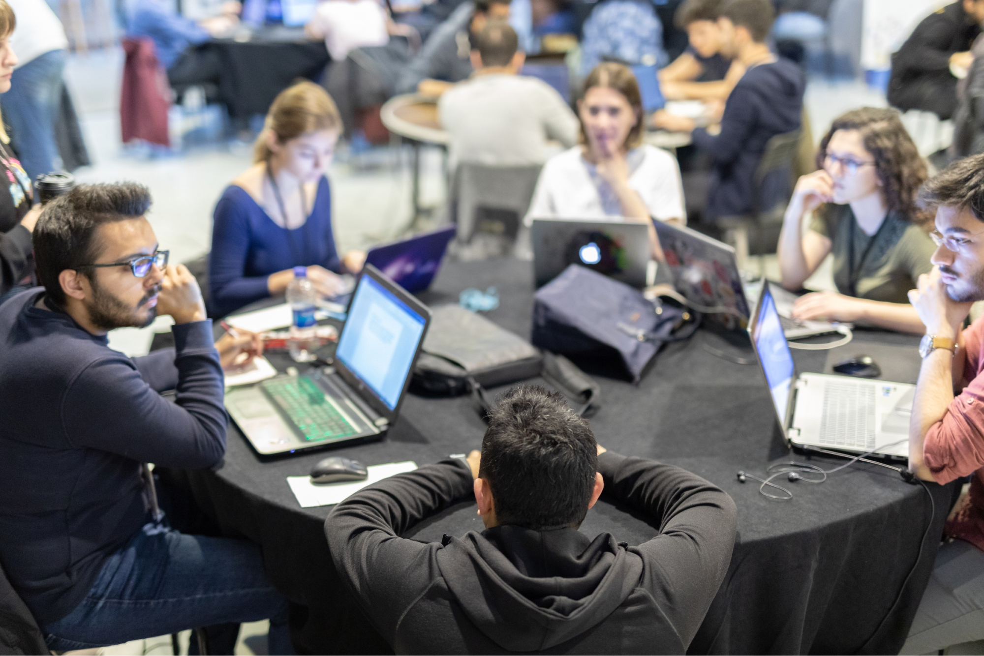 A group of people sitting at a round table with a black cloth, thinking, and working on their laptops.