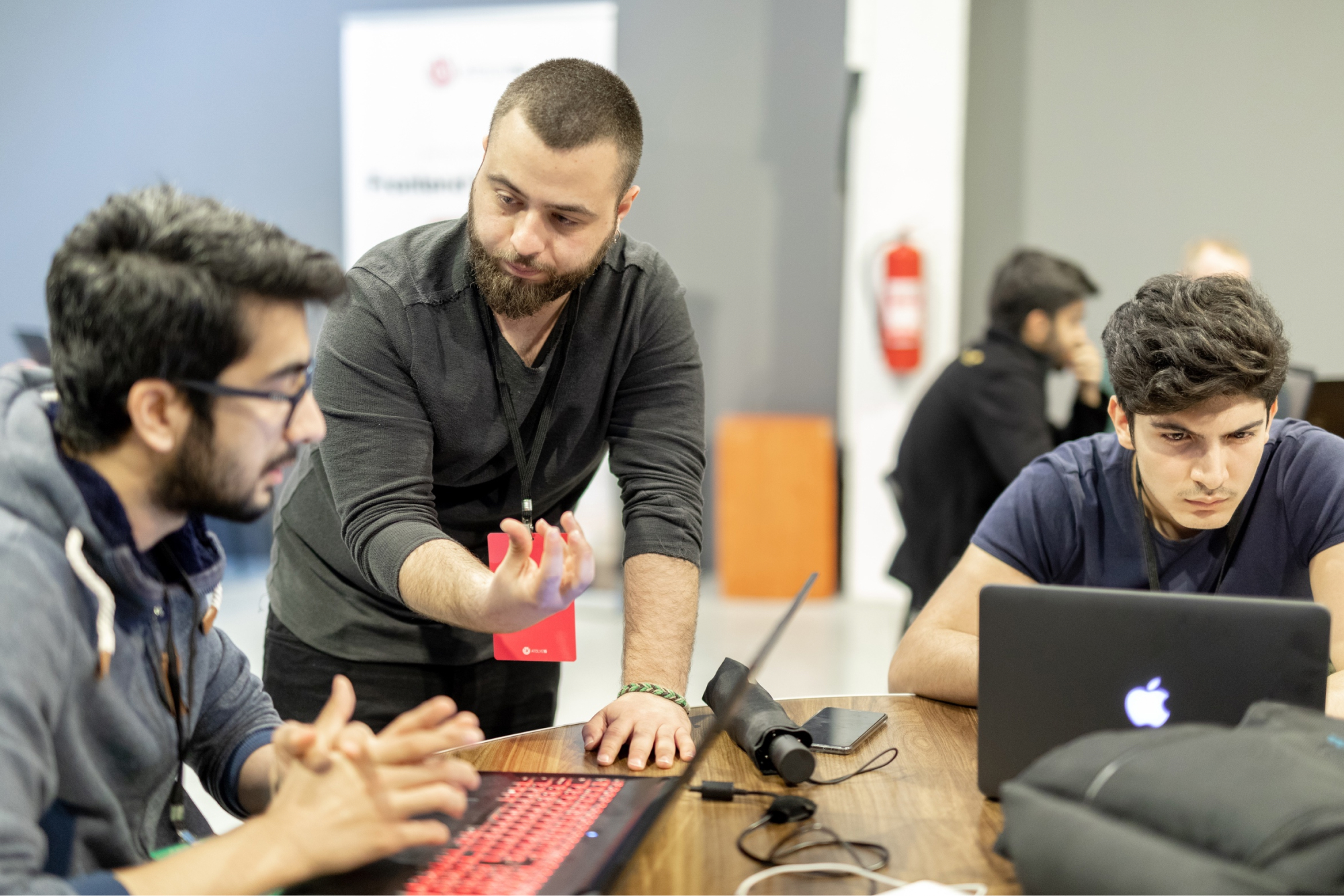 Two young boys working on their laptops and a man leaning against the table telling something with a hand gesture.