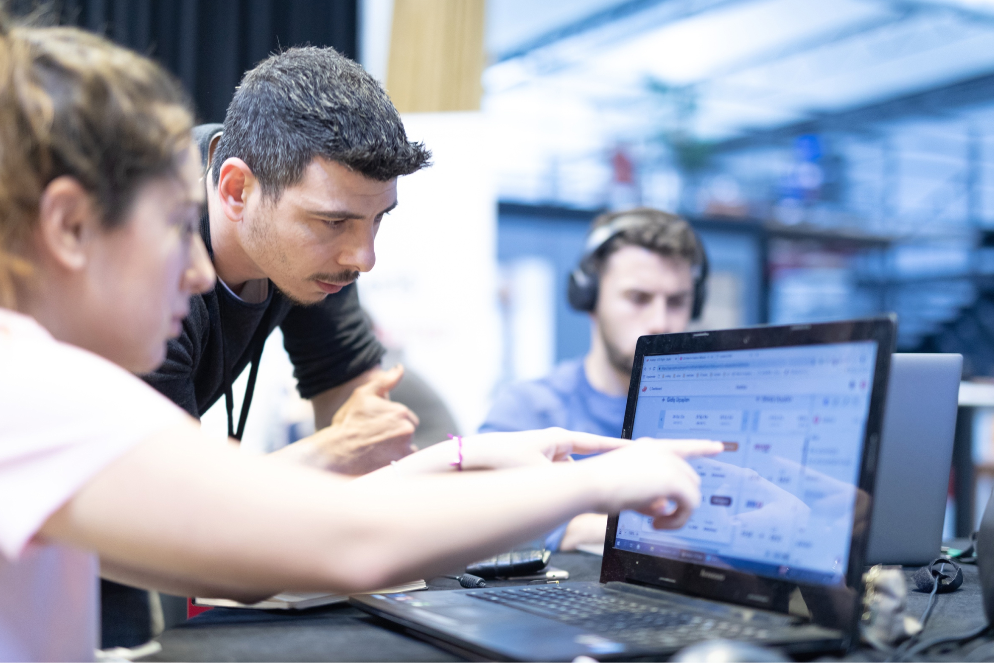 A man and a woman in a pink t-shirt pointing at a laptop screen and another wearing headphones while working.