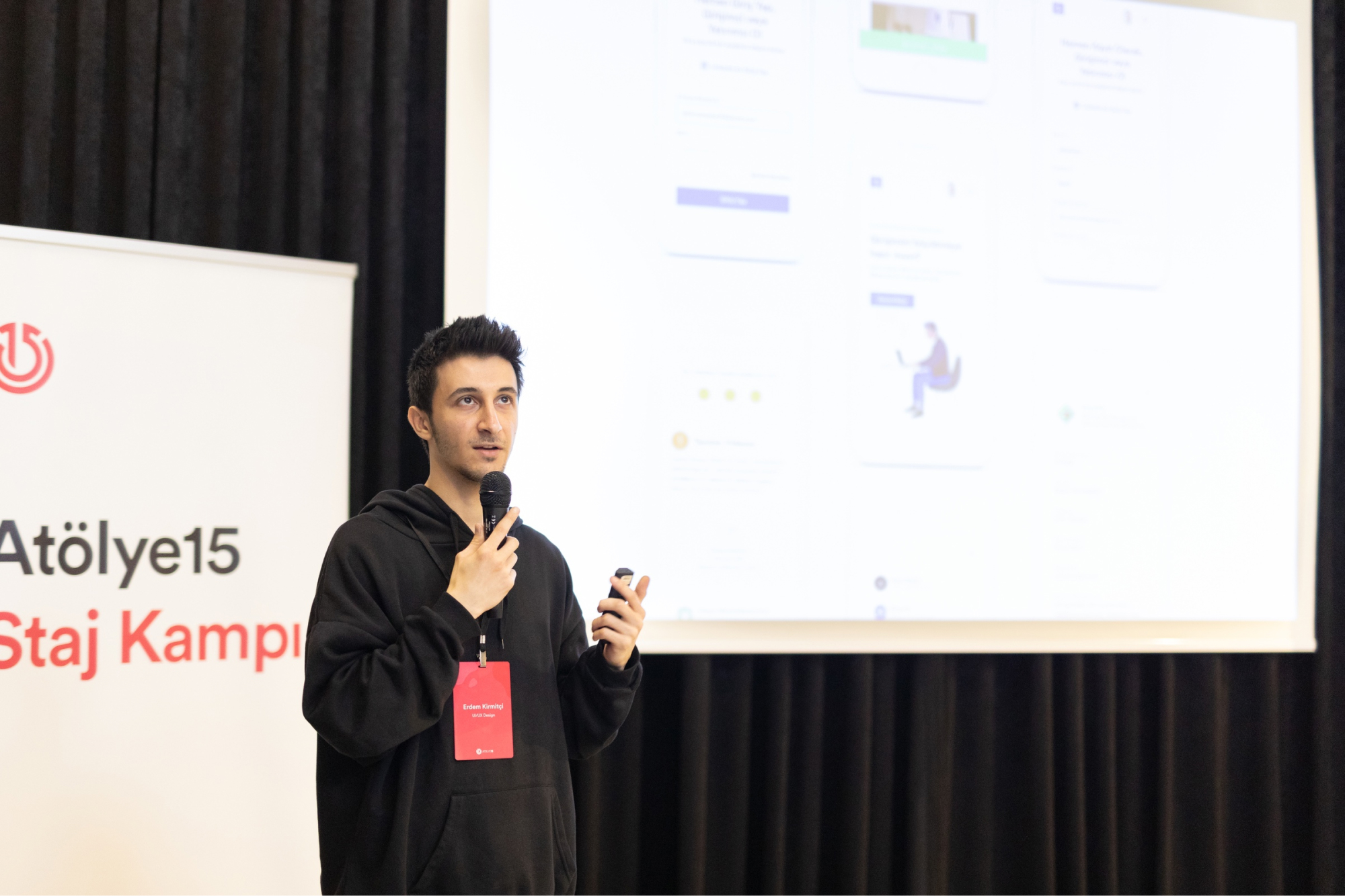 A man in a black hoodie and holdind a microhone making a speech in front of a projector and an “Atölye15 Staj Kampı” banner.