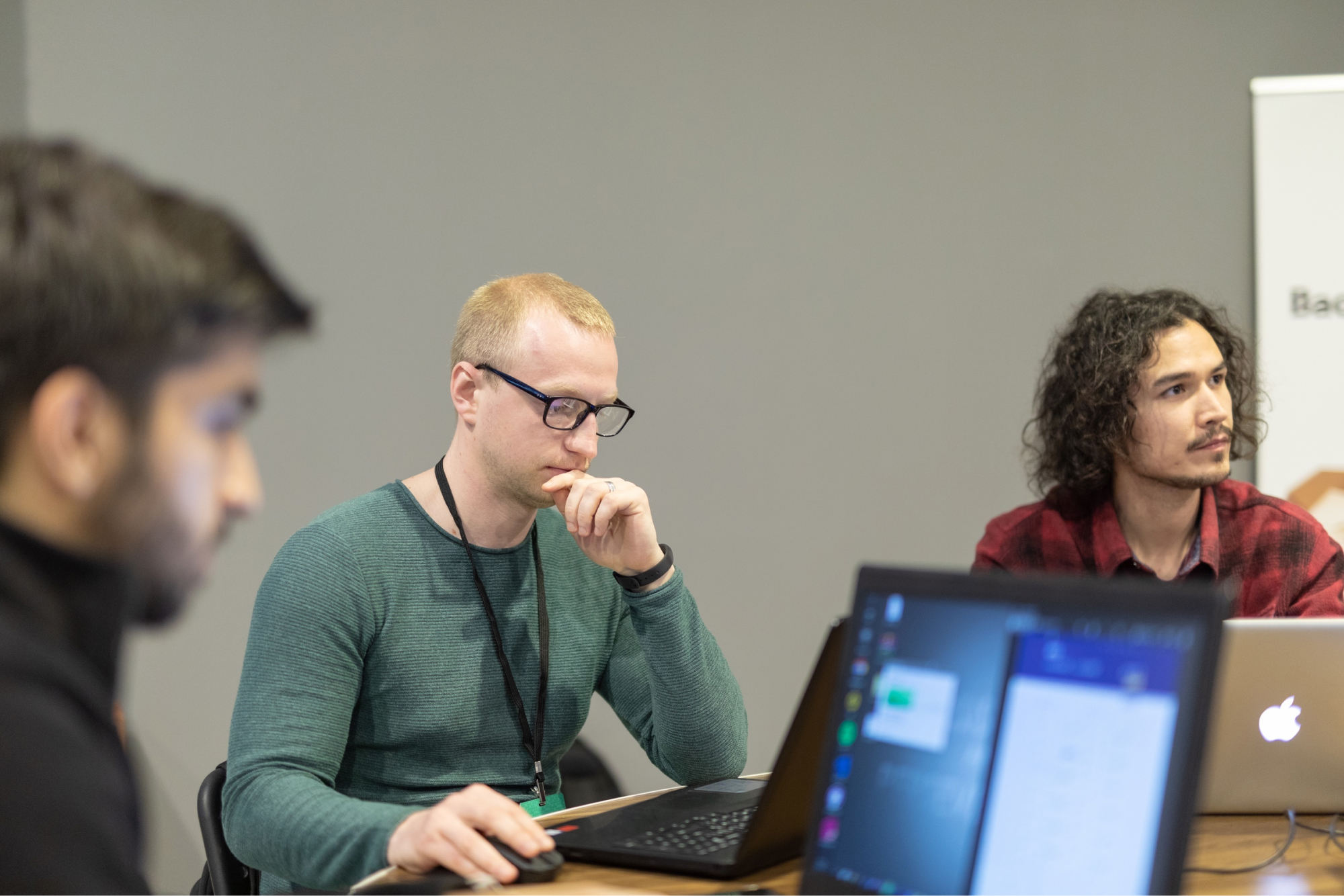 A ginger man in a green sweatshirt wearing glasses and next to a curly-haired man in a red plaid shirt working on their laptops.