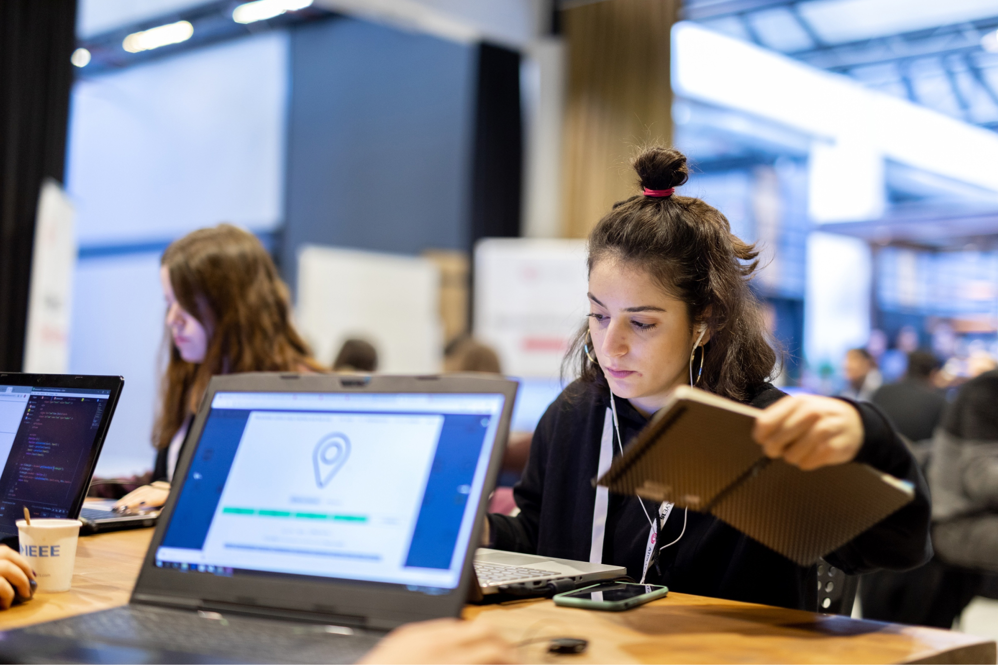 A girl with a bun in a black sweatshirt, sitting in front of a laptop screen showing a drop pin icon and holding a notebook.