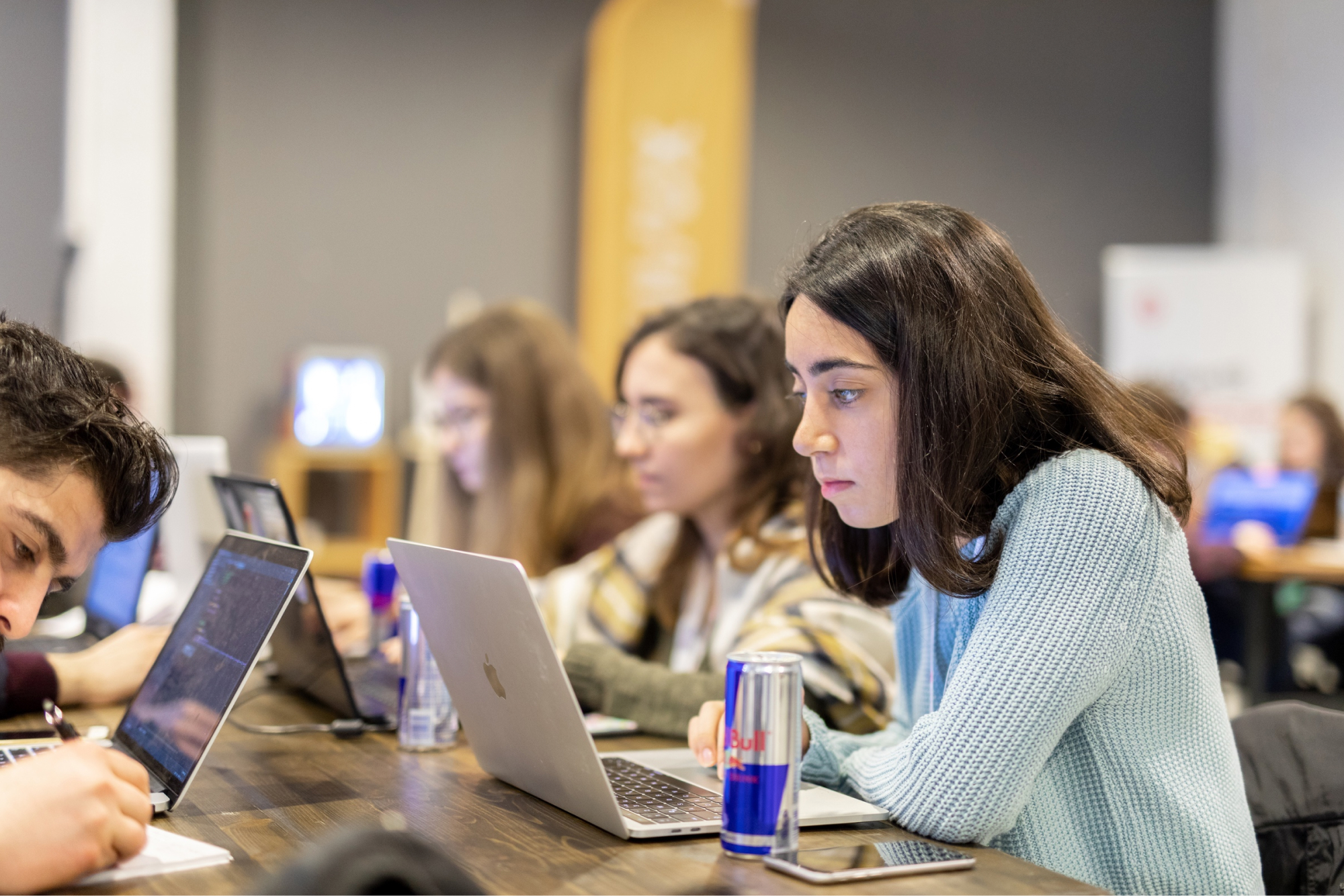 A girl in a green sweater sitting next to two other girls, looking at her MacBook that’s standing next to a can of Red Bull.