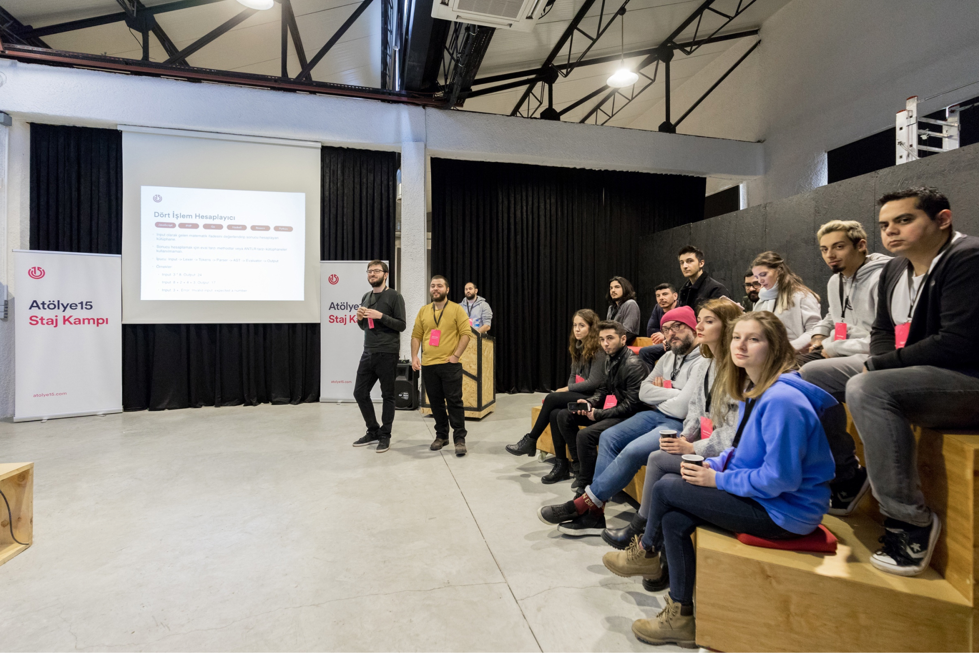A group of people, sitting on a wooden bench next to a presentation screen and an “Atölye15 Staj Kampı” banner.
