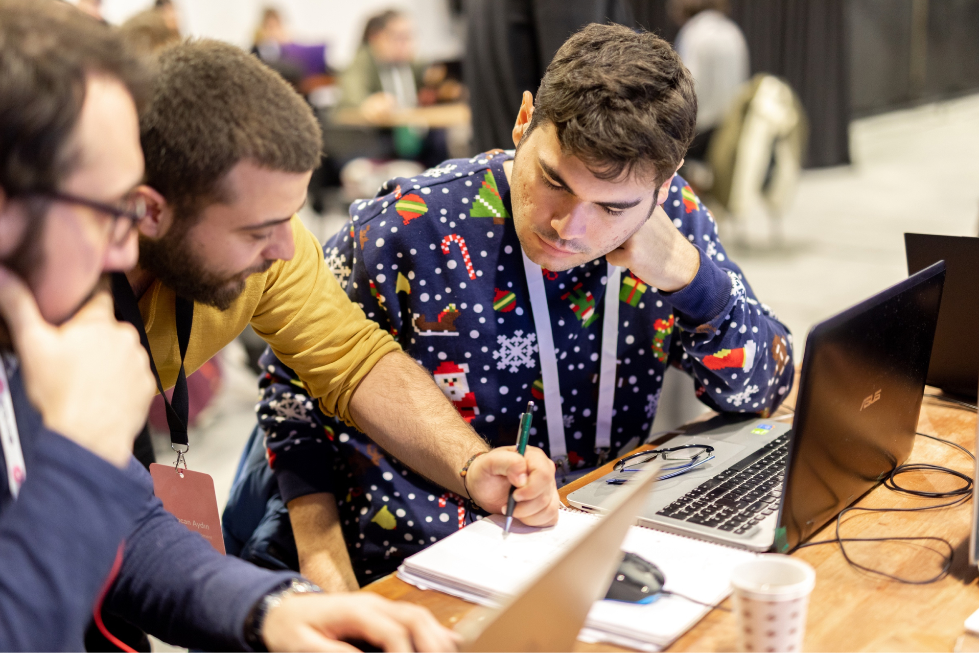 A man in a festive Christmas sweater sitting in front of a laptop and a man in a yellow sweatshirt writing with a green pen.