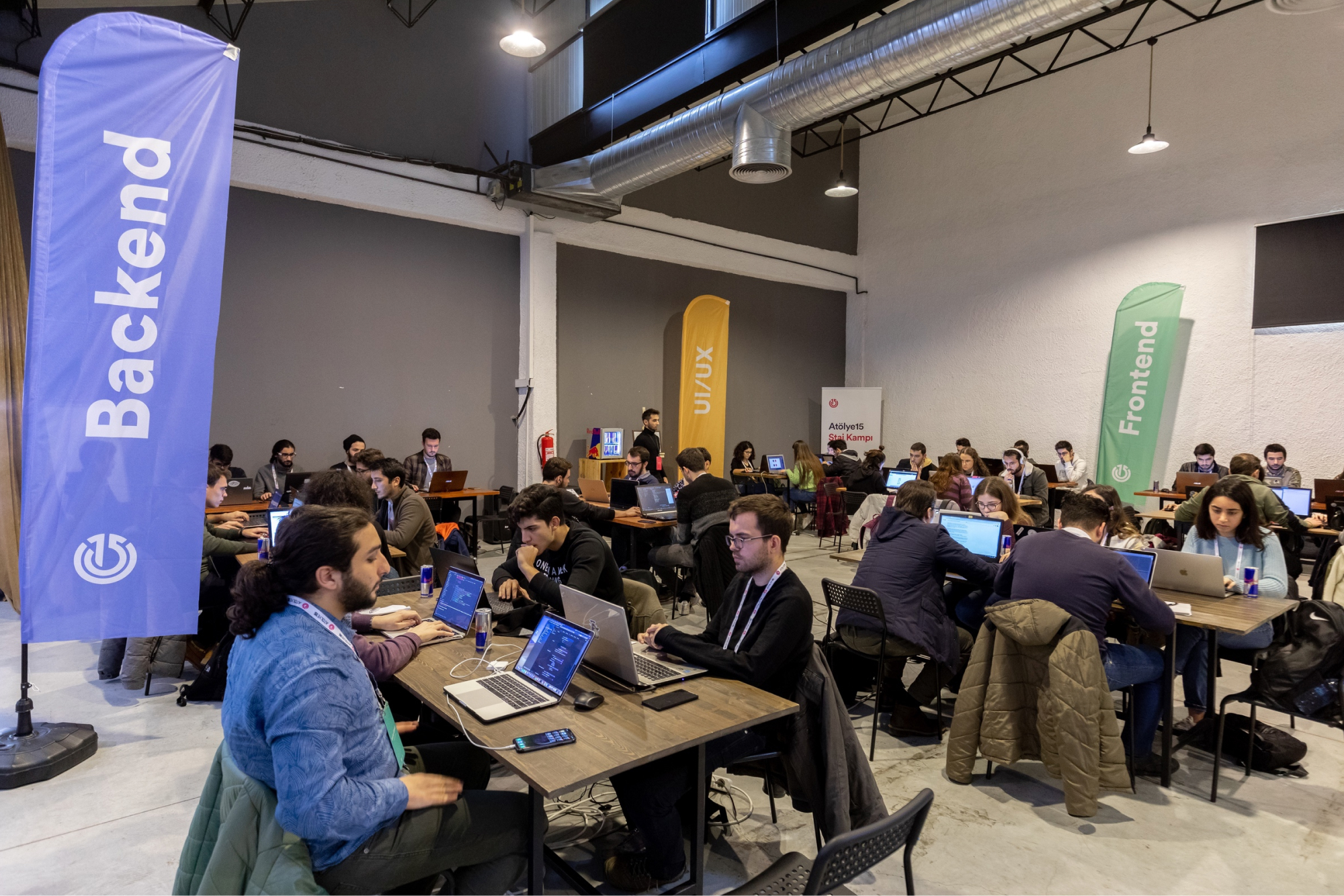 People sitting at tables, working on their laptops next two backend, frontend, UI/UX banners at the Atolye15 Internship Bootcamp.