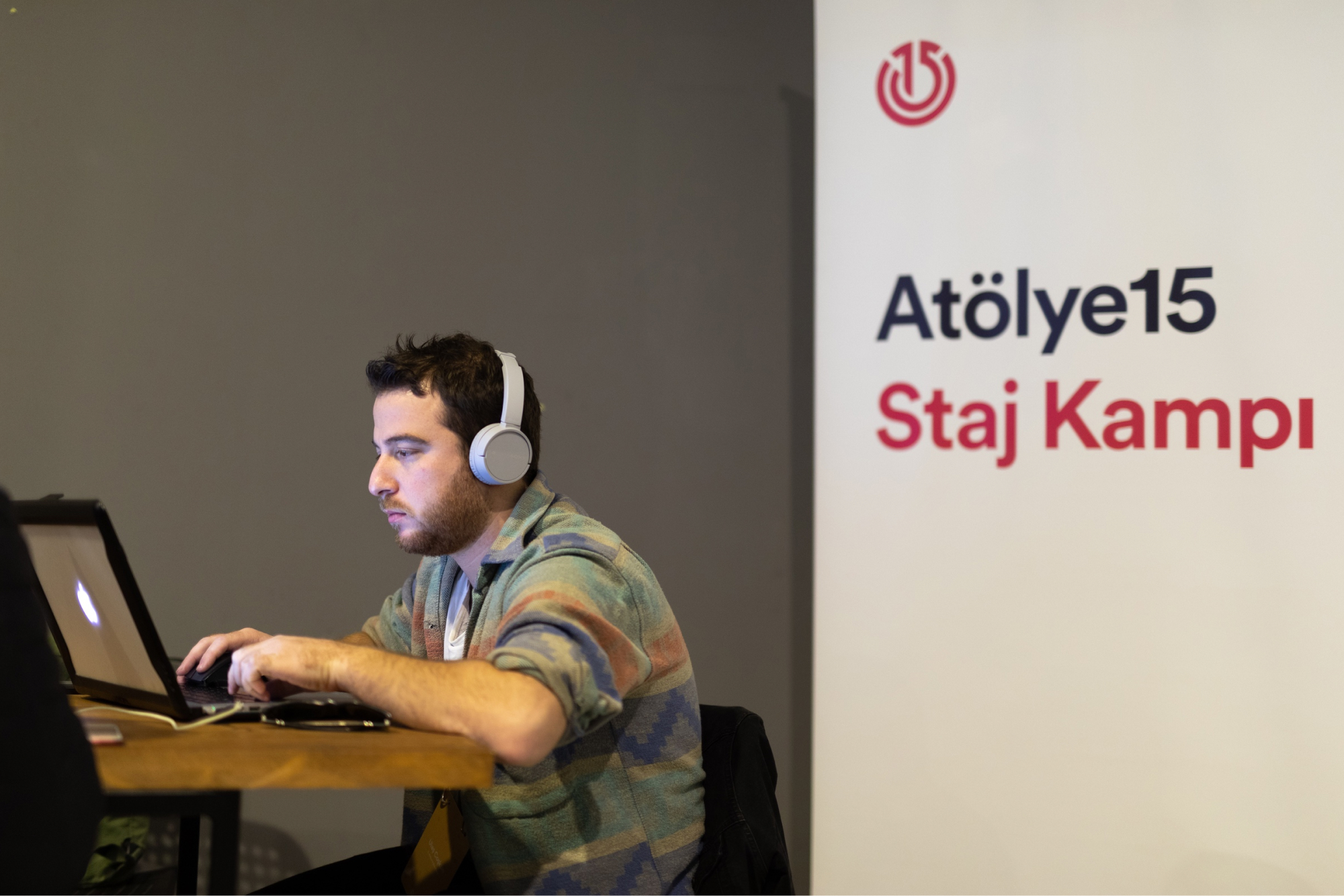 A man with white headphones sitting at a table and working on a MacBook in front of a banner that says “Atölye15 Staj Kampı.”