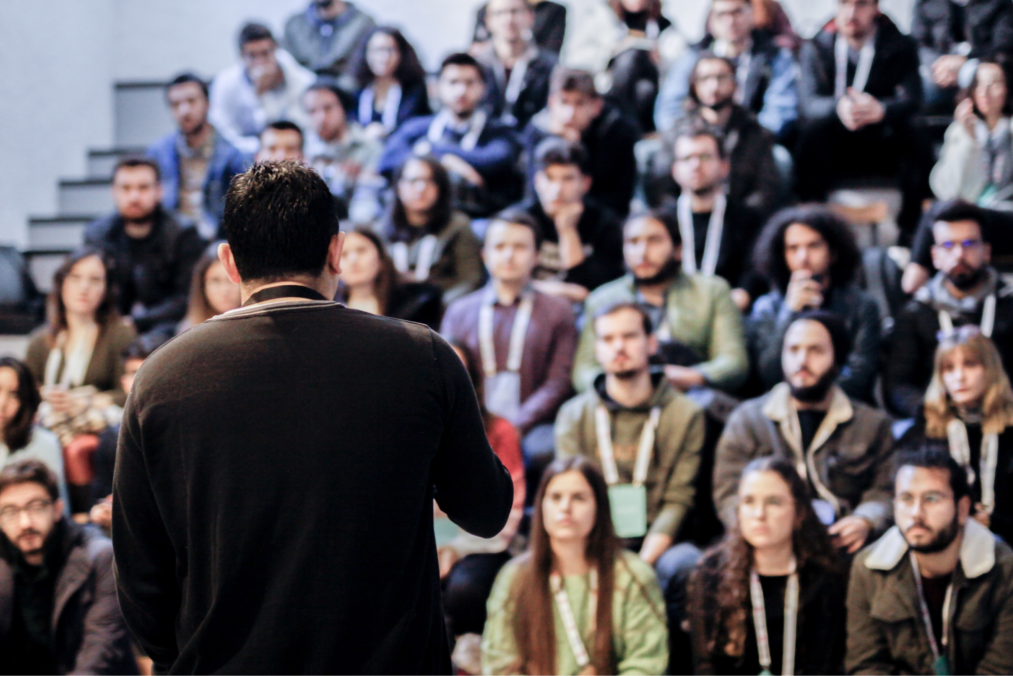 The back of a man wearing a black sweatshirt, talking to a group of people listening and sitting in a lecture hall.