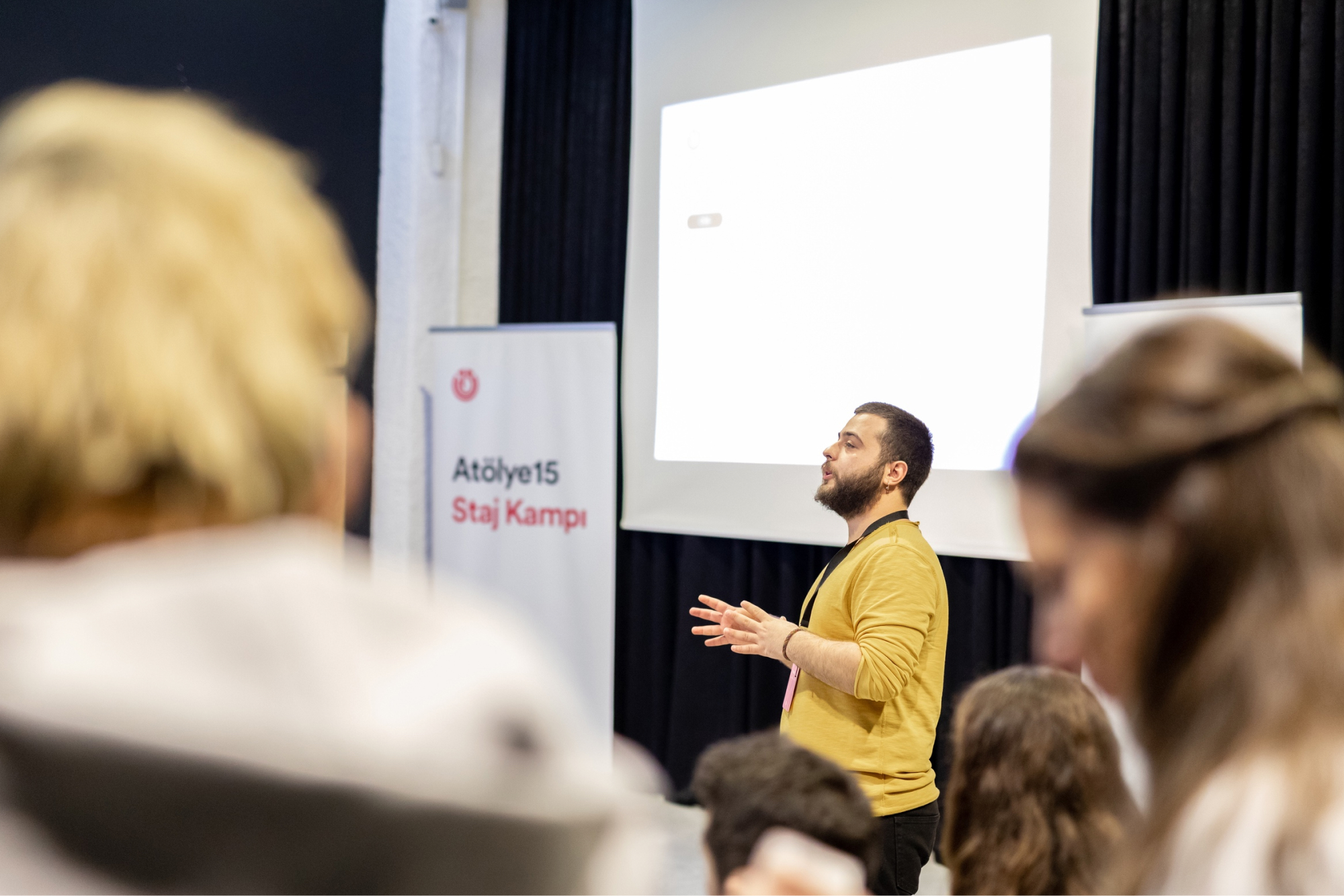 A man in a yellow sweatshirt, giving a presentation on stage next to a white banner that says “Atölye15 Staj Kampı.”