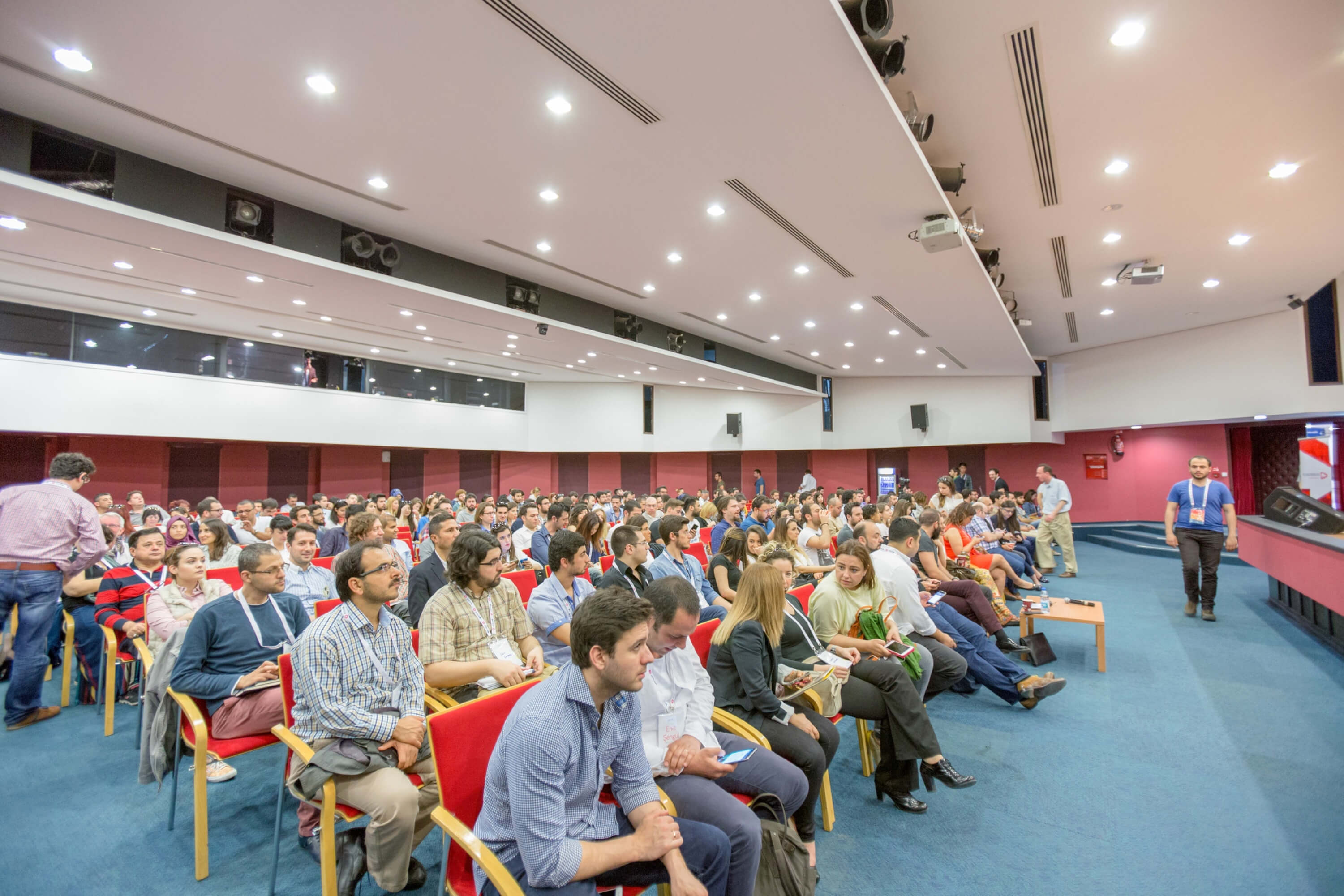 A group of people sitting in red chairs on a blue blanket under spotlights in a conference hall for IzmirUP.