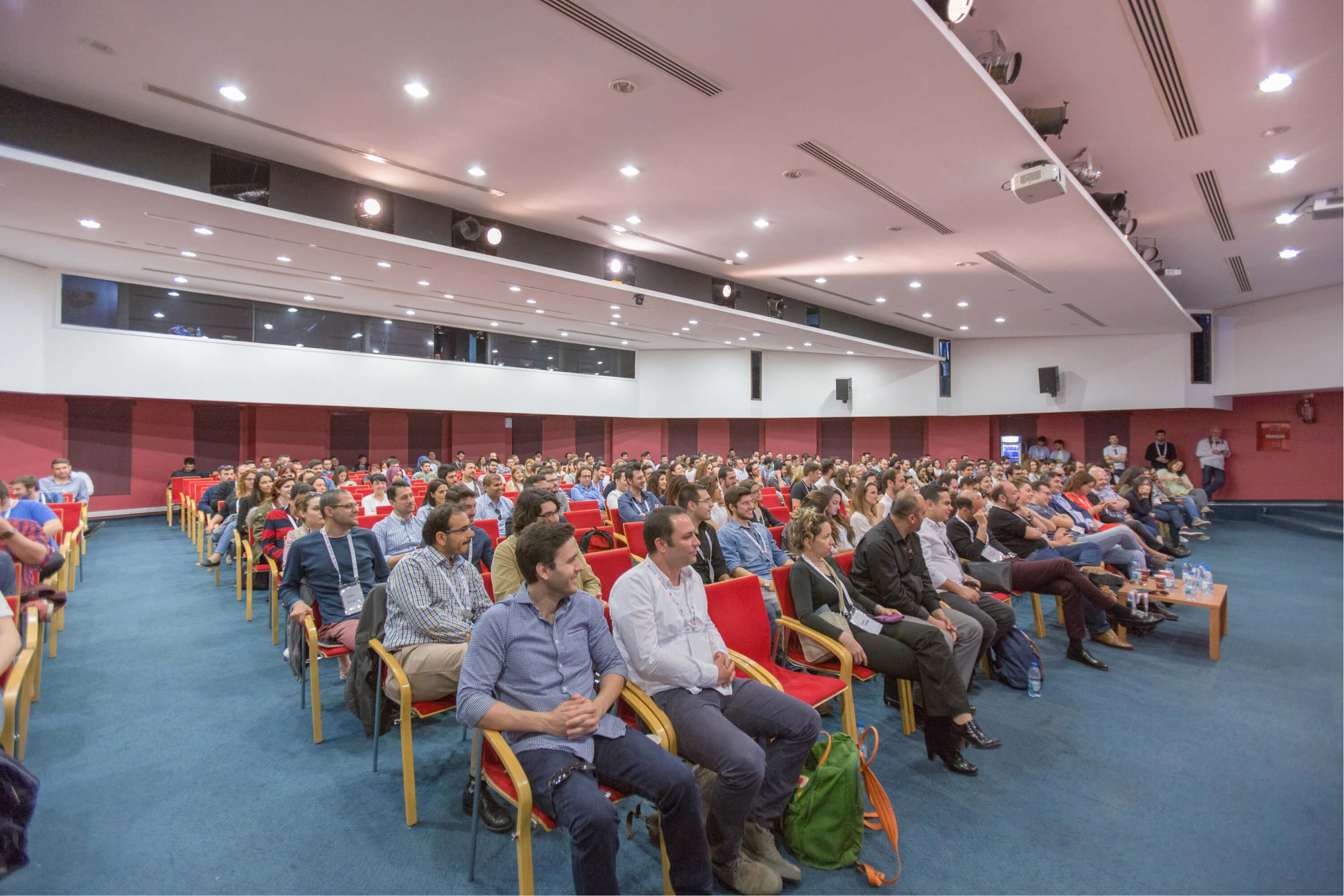 A group of people sitting in red chairs on a blue blanket under spotlights in a conference hall for IzmirUP.