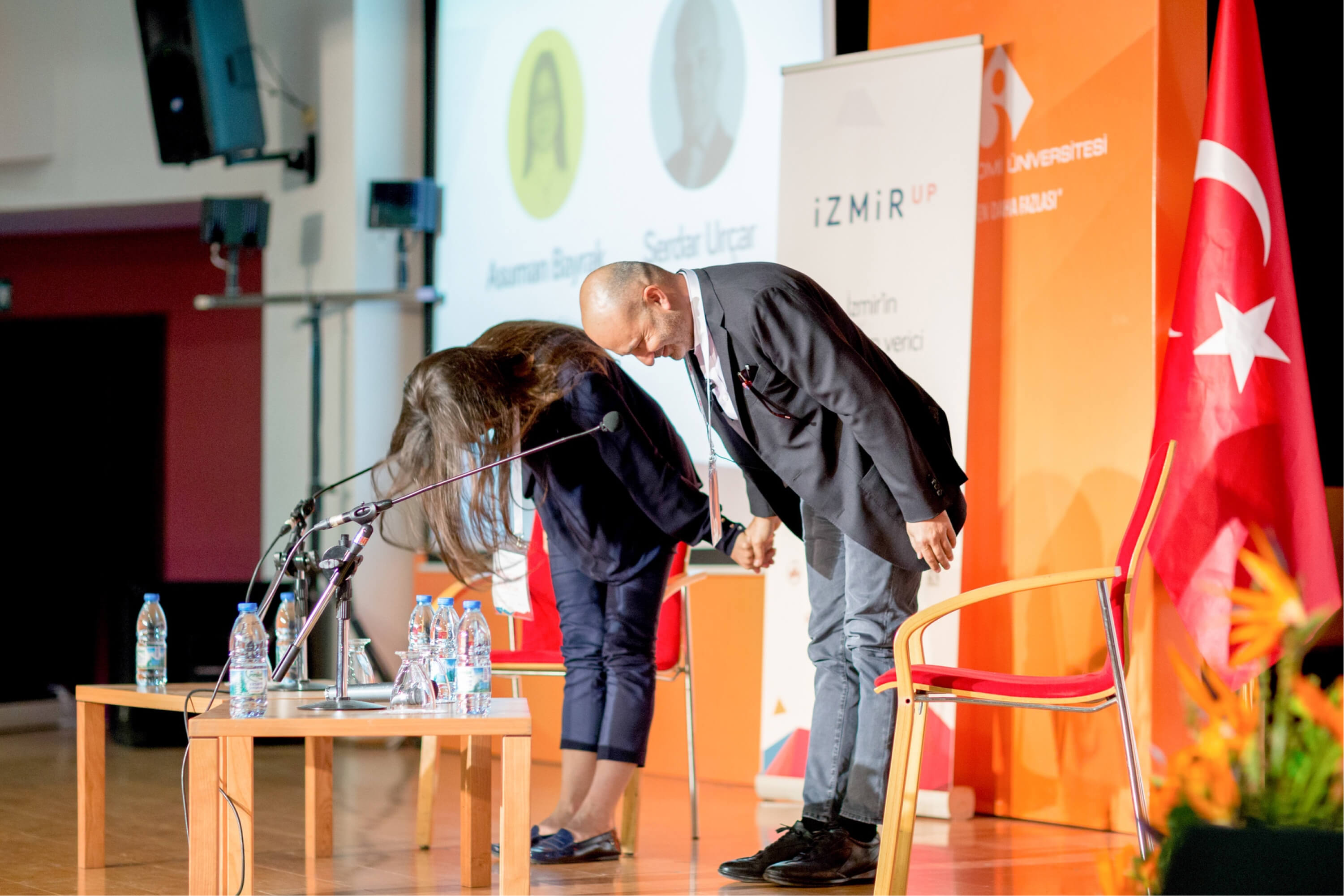 A long-haired woman and a bald man in a suit holding hands and taking a bow in front of the Turkish flag and an IzmirUP banner.