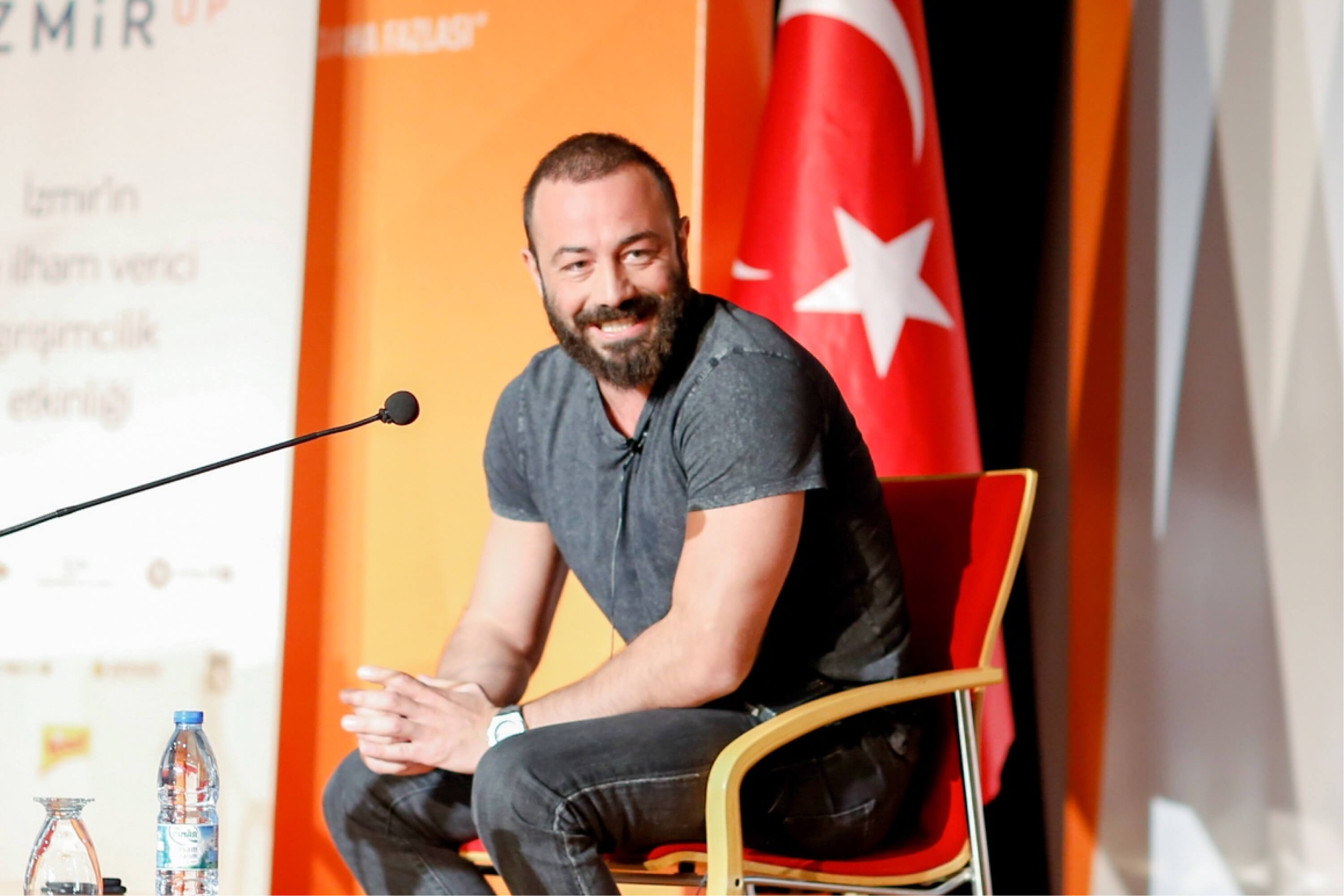 A bearded man in a grey t-shirt sitting in a red chair at IzmirUP in front of a Turkish flag, a water bottle, and a microphone.