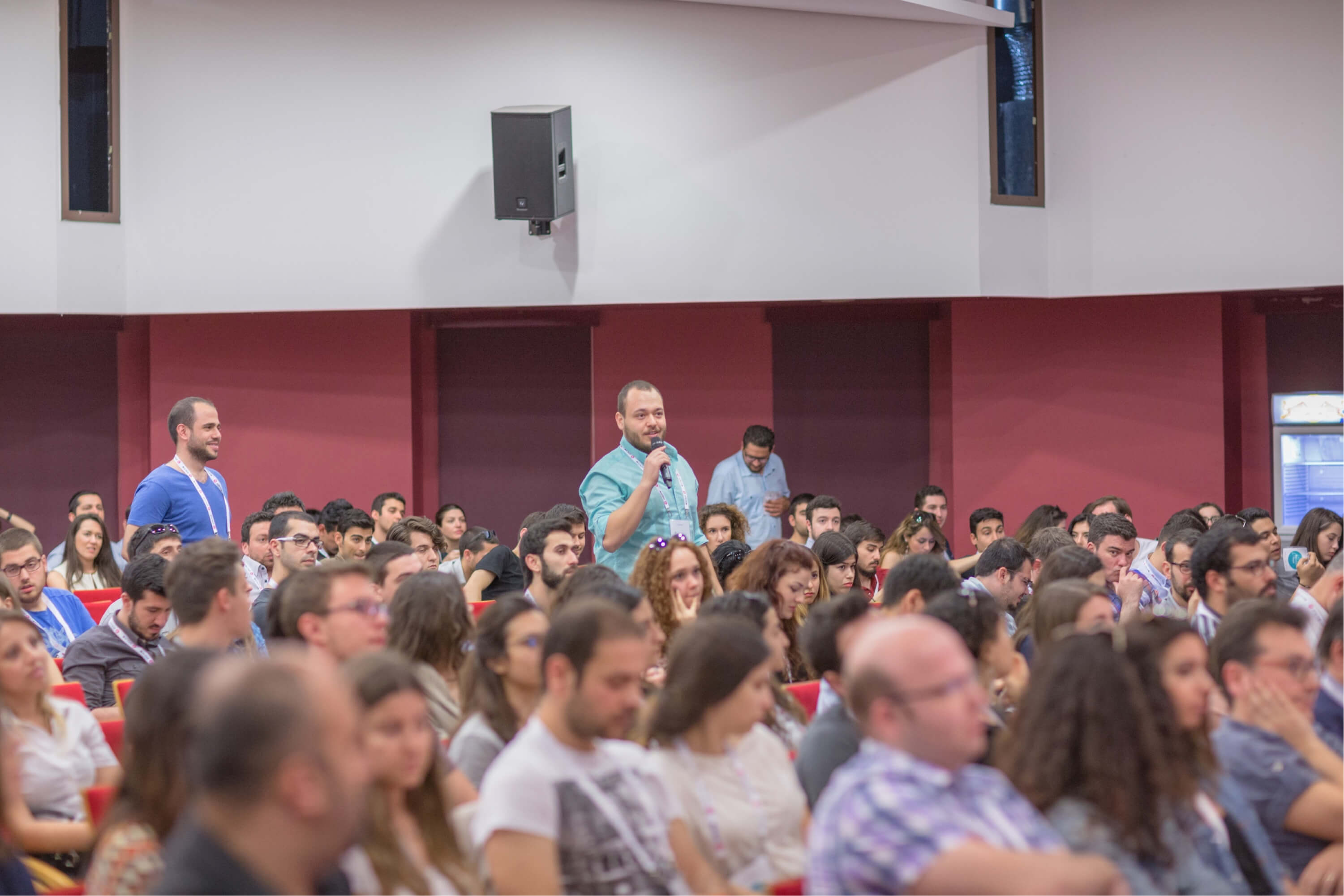 A man in a green shirt talking to a microphone while standing up in a crowd of people sitting in a conference hall.