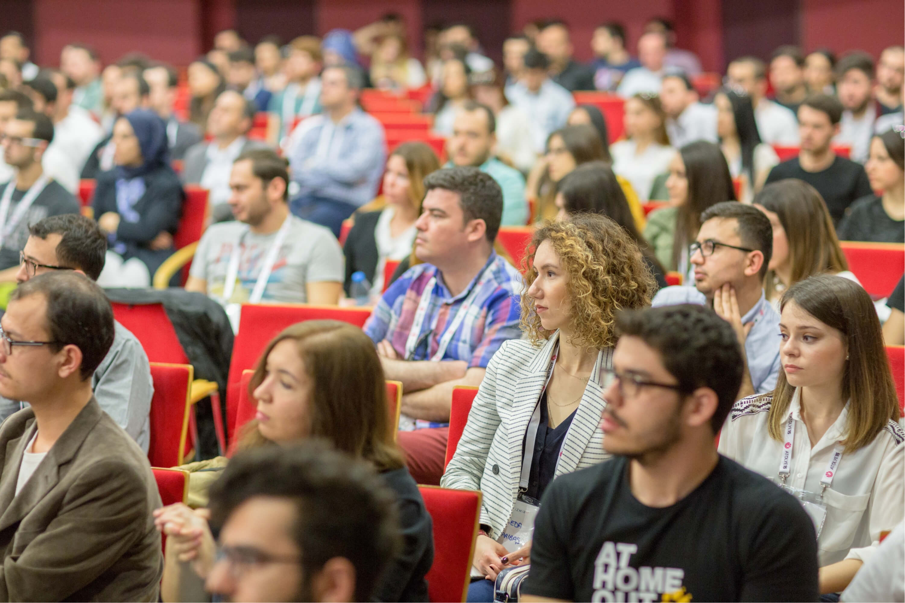 A crowded group of people sitting in red chairs in a conference hall and listening to a speech.
