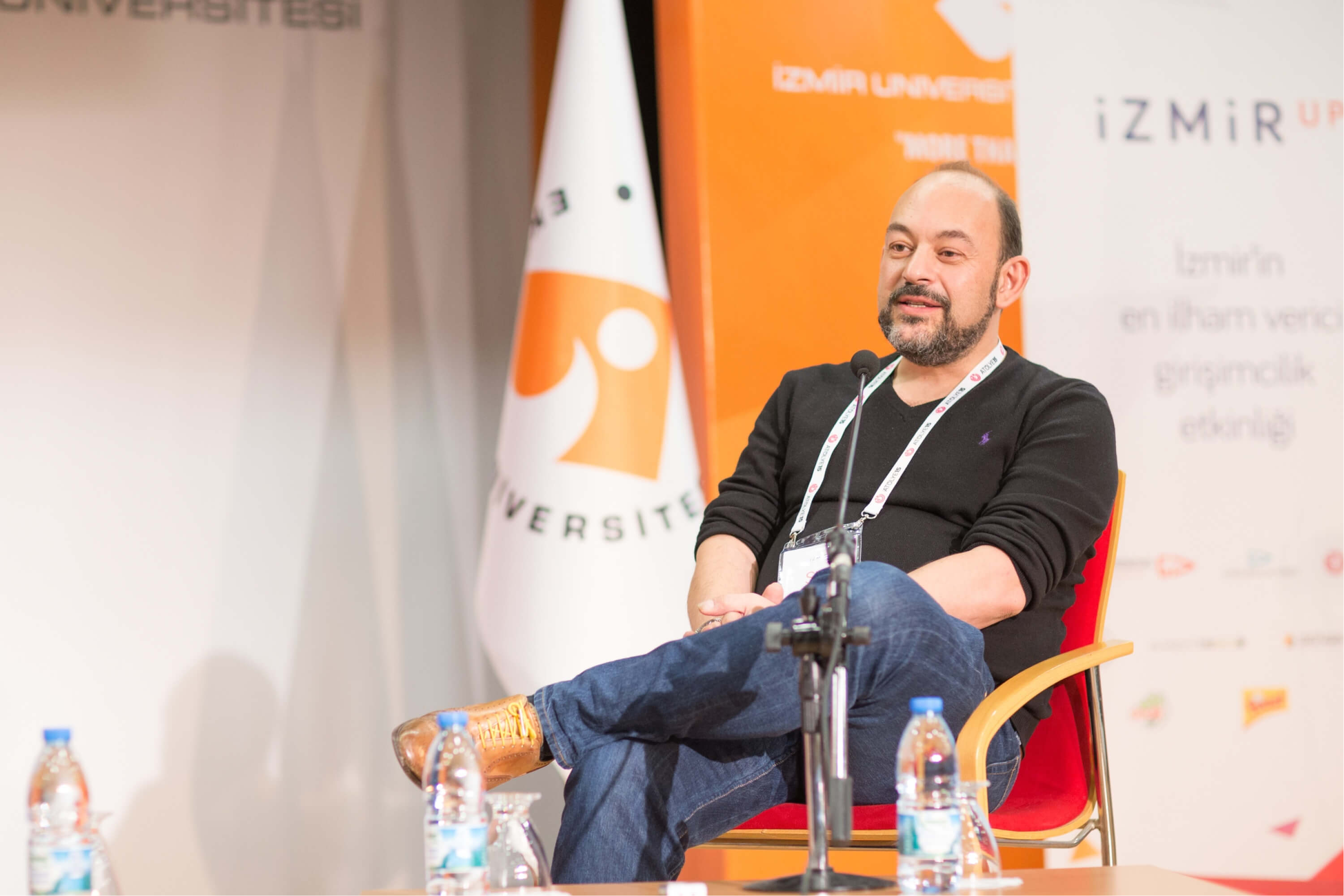 A middle-aged man in a black sweatshirt, sitting in a chair in front of a microphone and a banner that says “IzmirUP.”