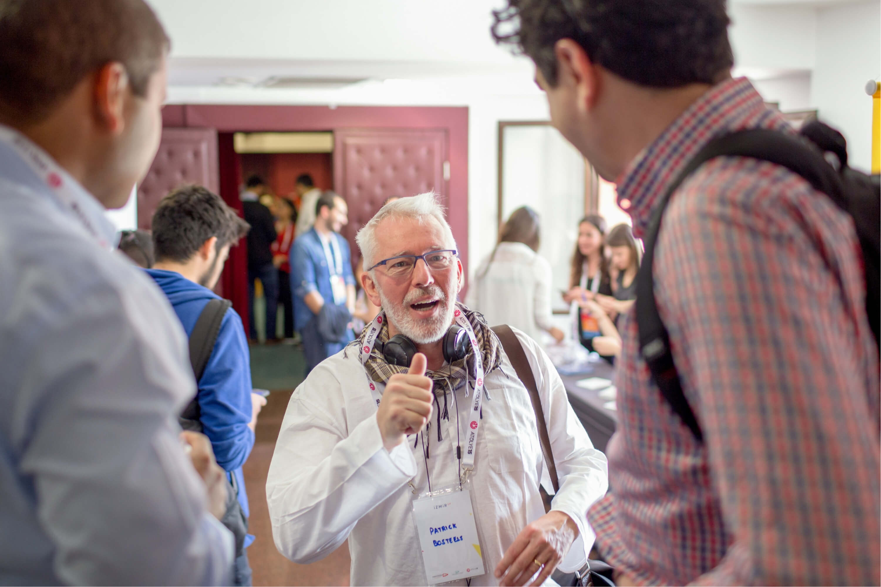 An old man in a white shirt, wearing glasses, headphones, and a name tag that reads “Patrick Bosteels” talking to a man.