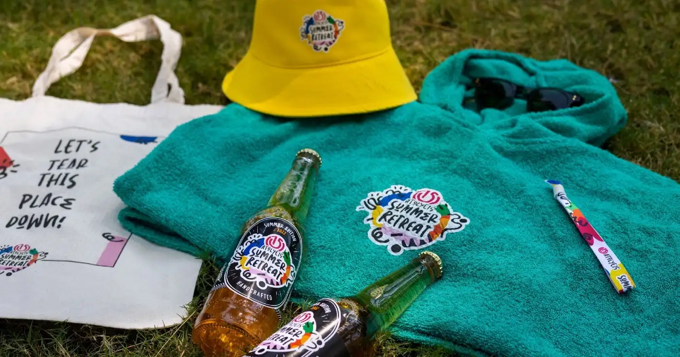 A green towel poncho, a yellow bucket hat, a tote bag, and two beer bottles from the Atolye15 Summer Retreat Kit.