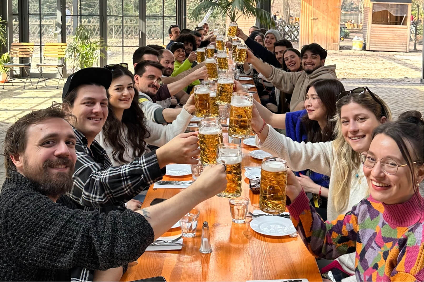 The Atolye15 Team sitting at a long wooden table, smiling and raising beer glasses in a Biergarten in Berlin.