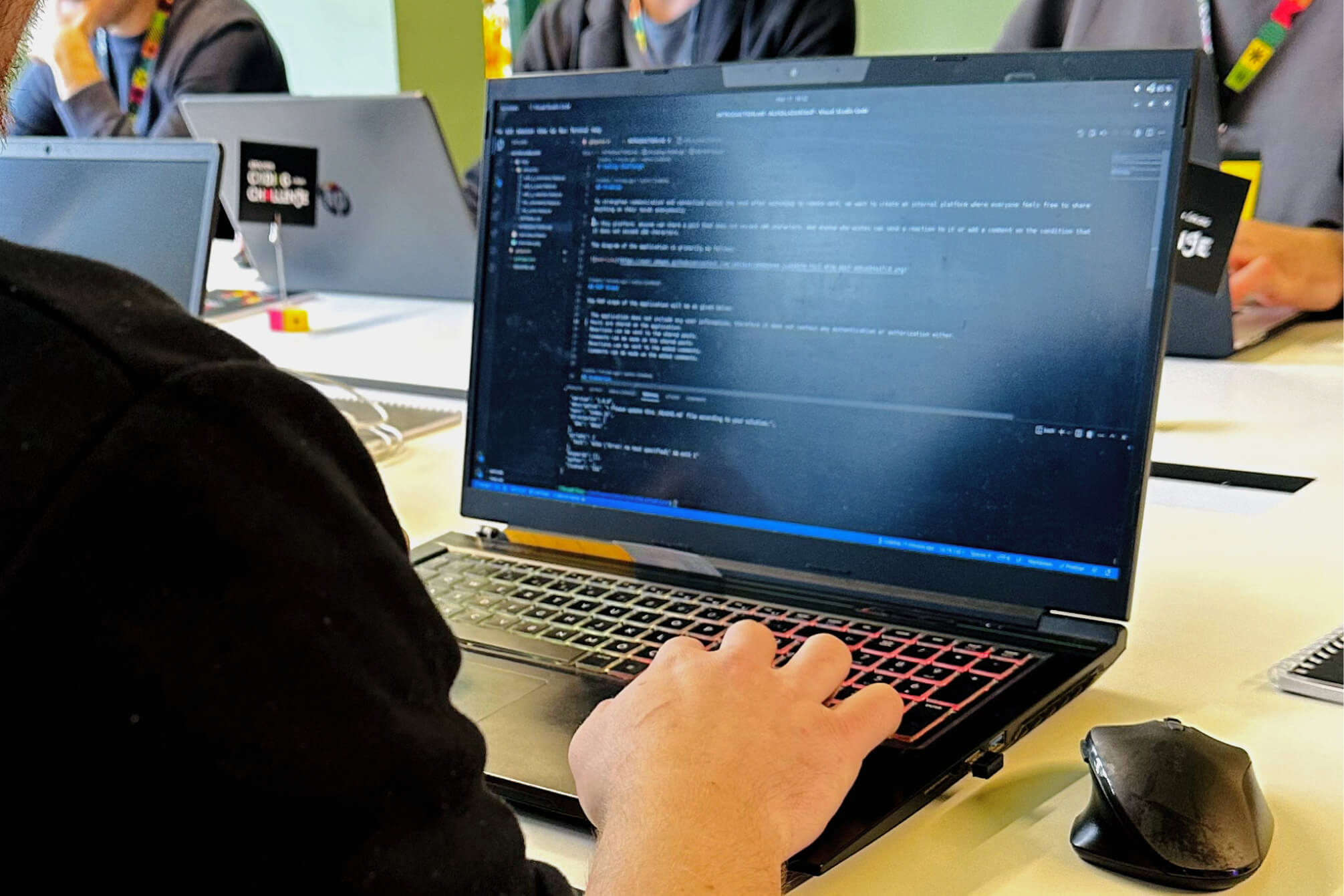 A hand on the keyboard of a black laptop and lines of code on the screen next to an Atolye15 Coding Challenge Ankara card.