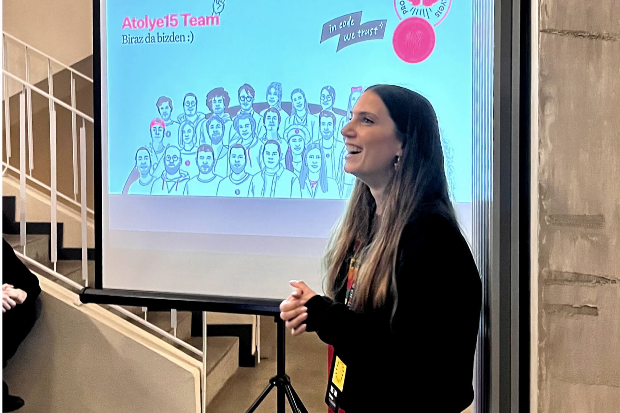 A blonde woman smiling and giving a presentation in front of a screen that shows an illustration of the Atolye15 Team members.
