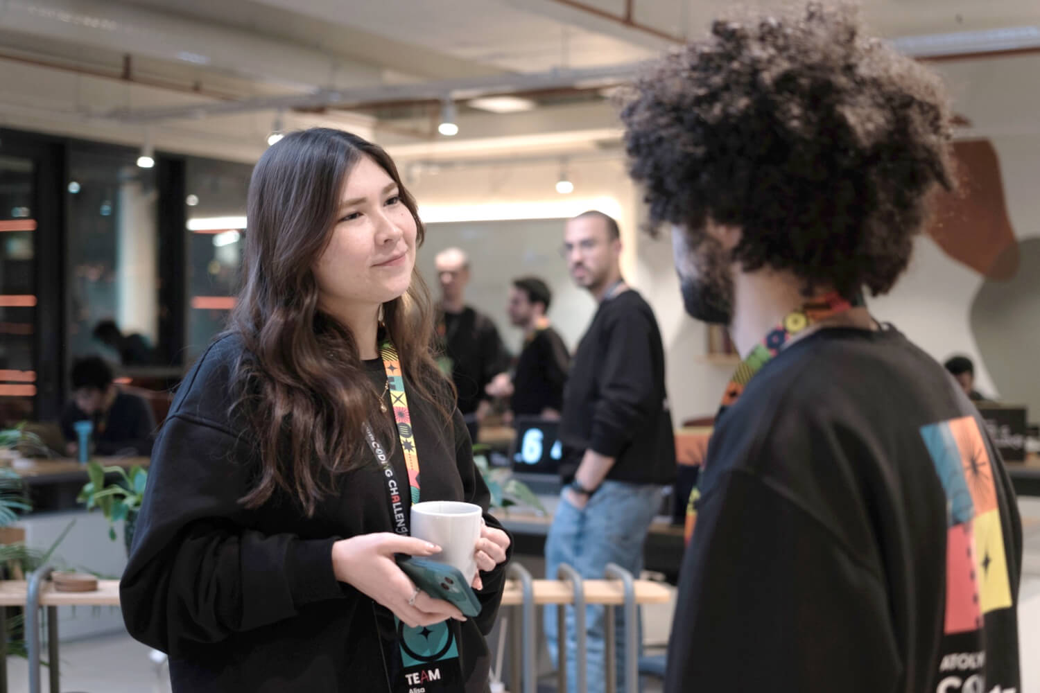 A young woman holding a white mug and talking to a curly-haired man who’s wearing a black Atolye15 Coding Challenge sweatshirt.