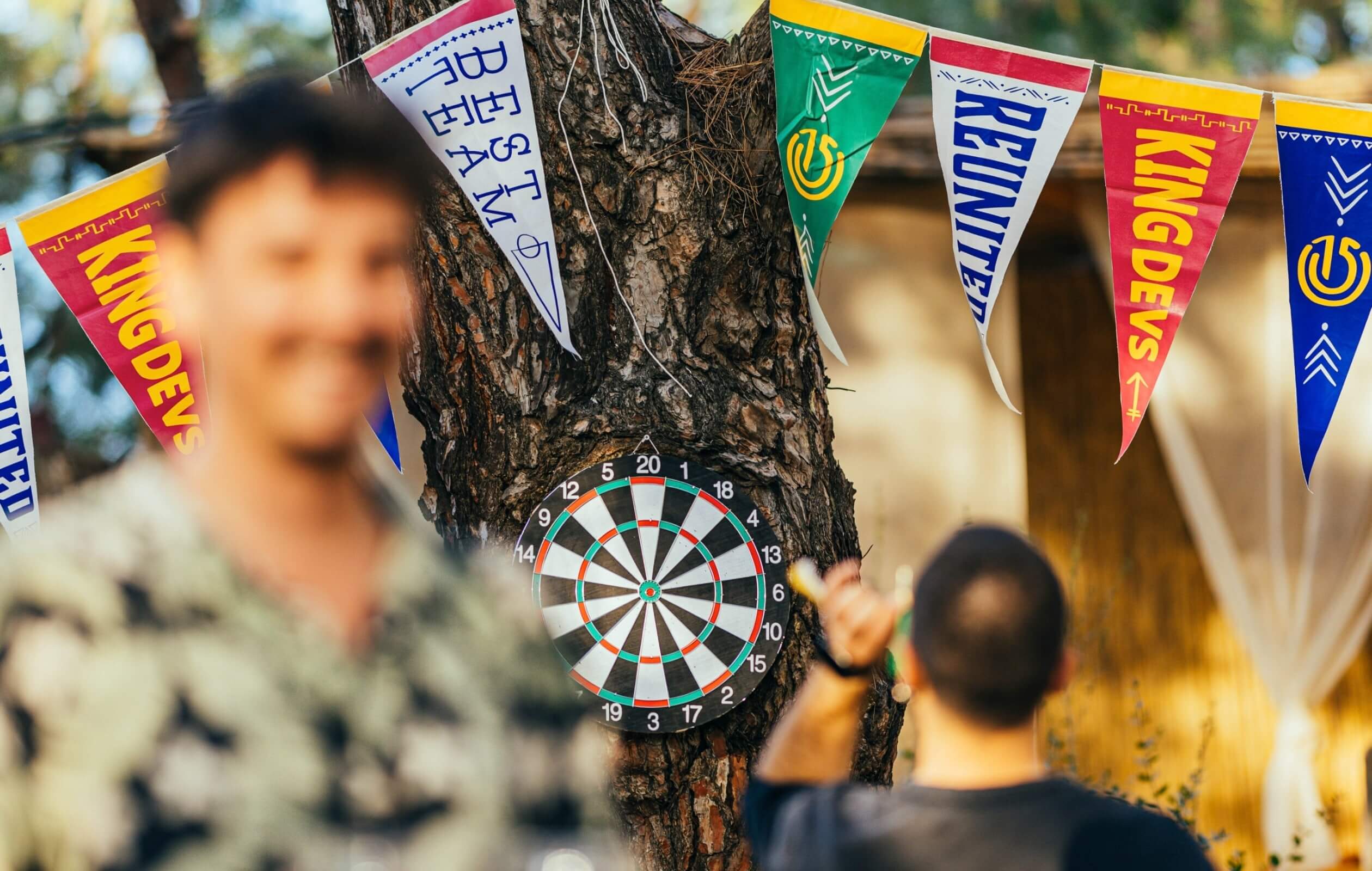 A silhouette of a smiling young man standing beside another individual playing darts, set against a tree decorated with Atolye15 Homecoming flags.