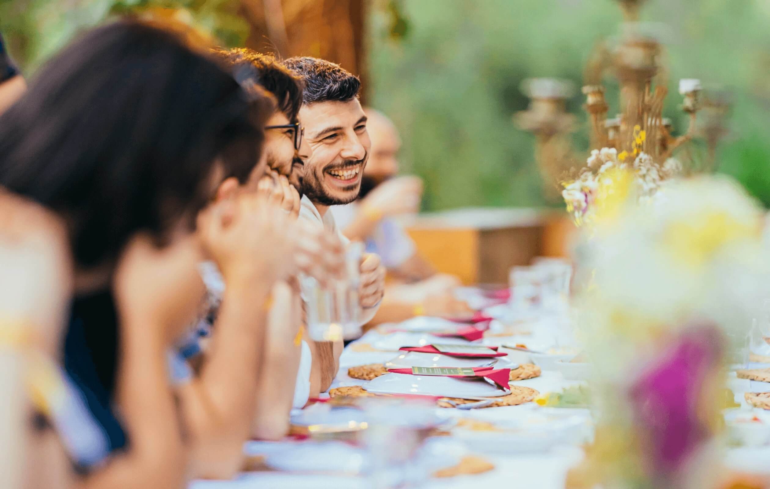 A line of people seated at a long dining table, with the camera focus on a man who is smiling.