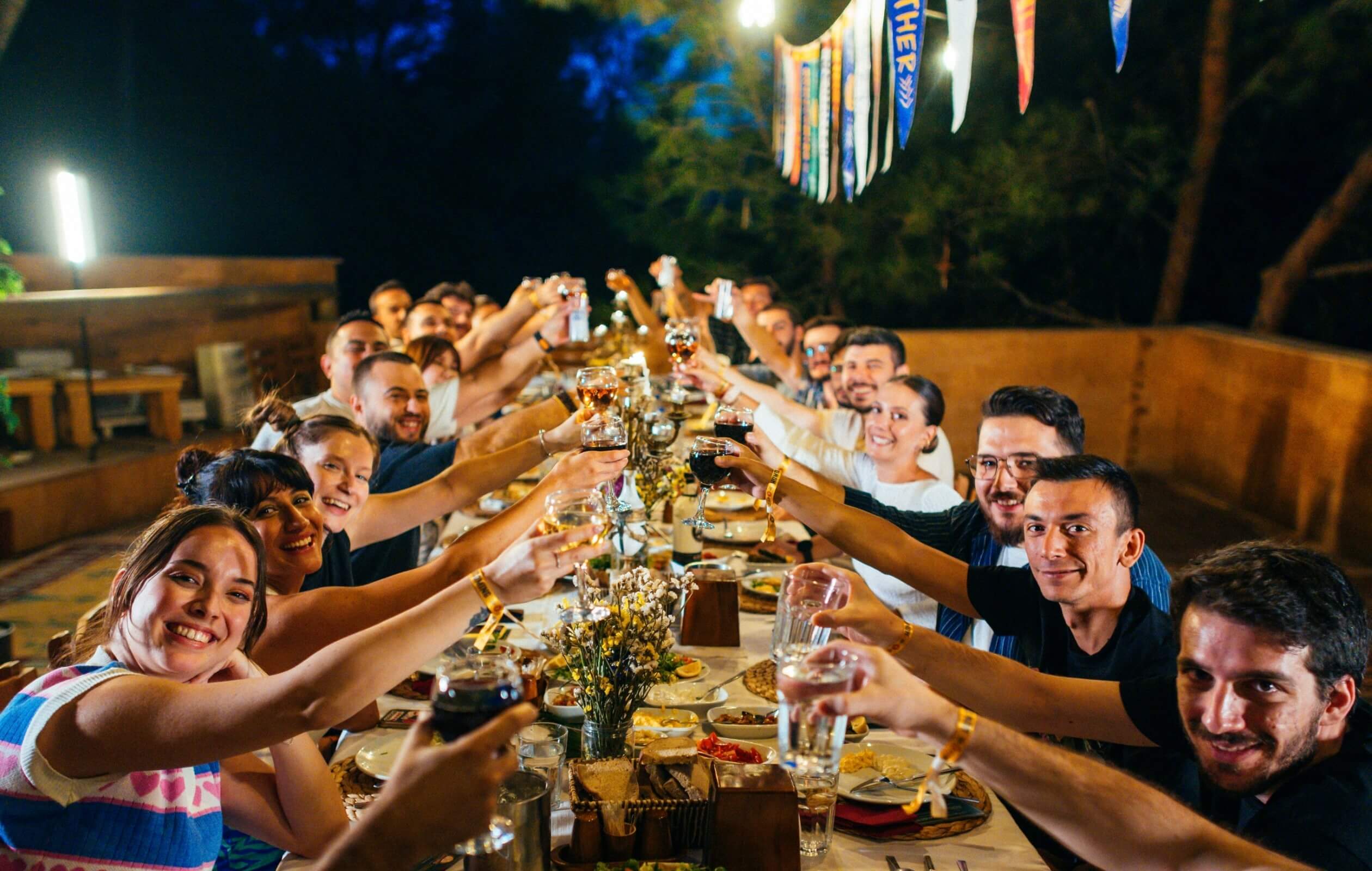 A group of people seated at a long dining table, raising their drinks, and clinking their glasses together.