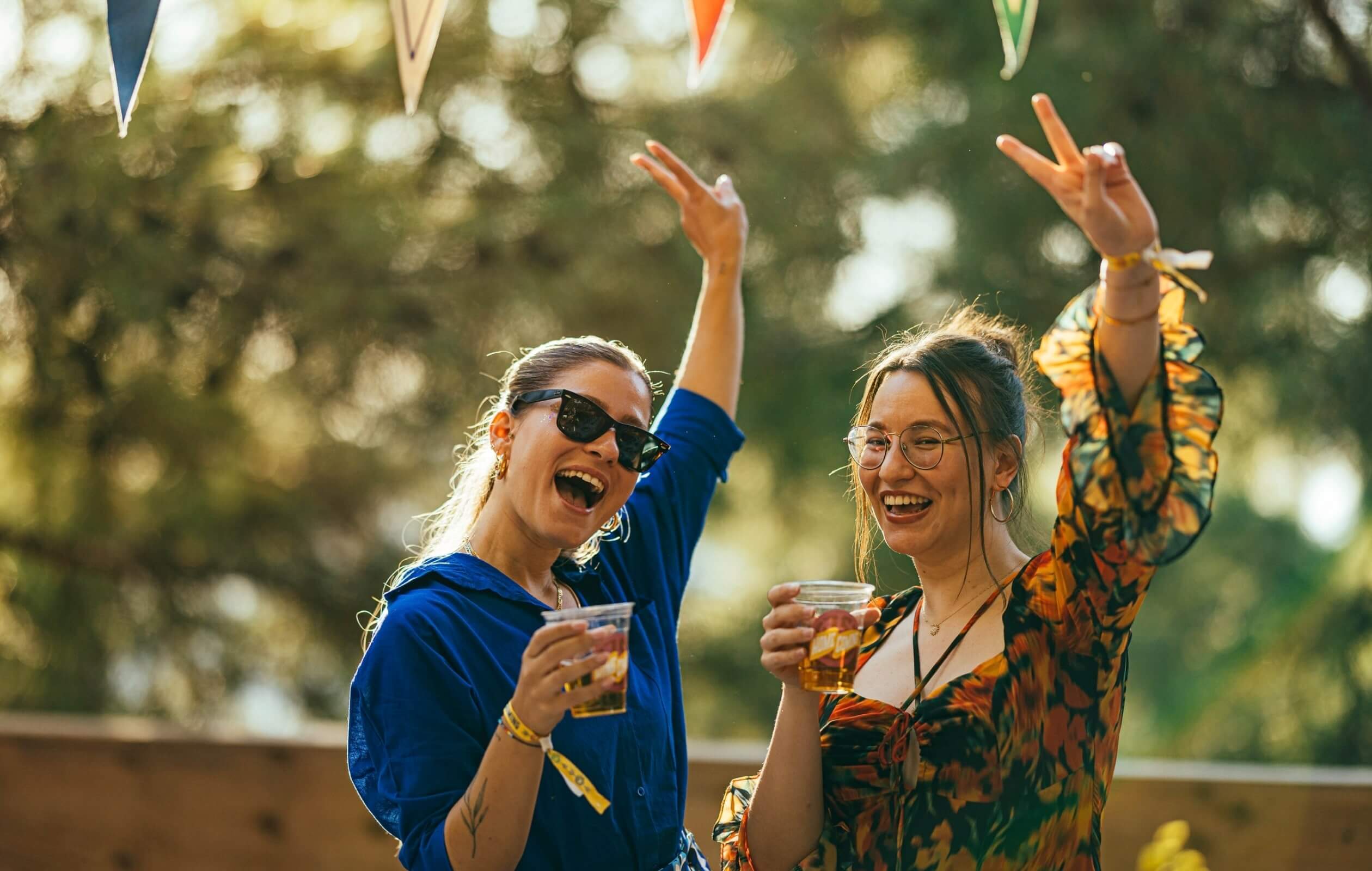 Two young women posing together, flashing peace signs with their hands raised in the air, and holding beer glasses that feature the Atolye15 Homecoming logo.