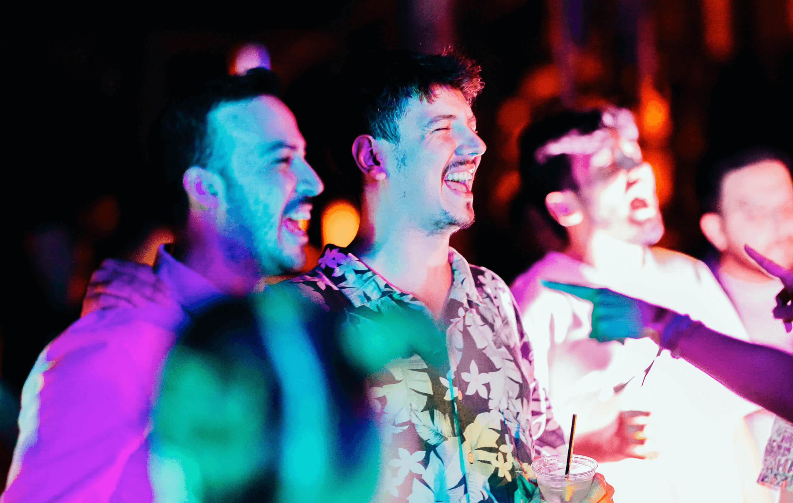 Three young men dancing and singing at a concert, illuminated by colorful lights and holding cocktails.