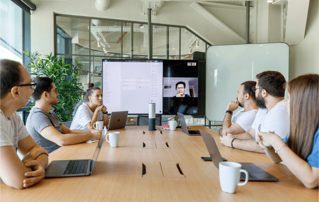 Team in a conference room attentively watching a coworker on a video call, with laptops and coffee on table.