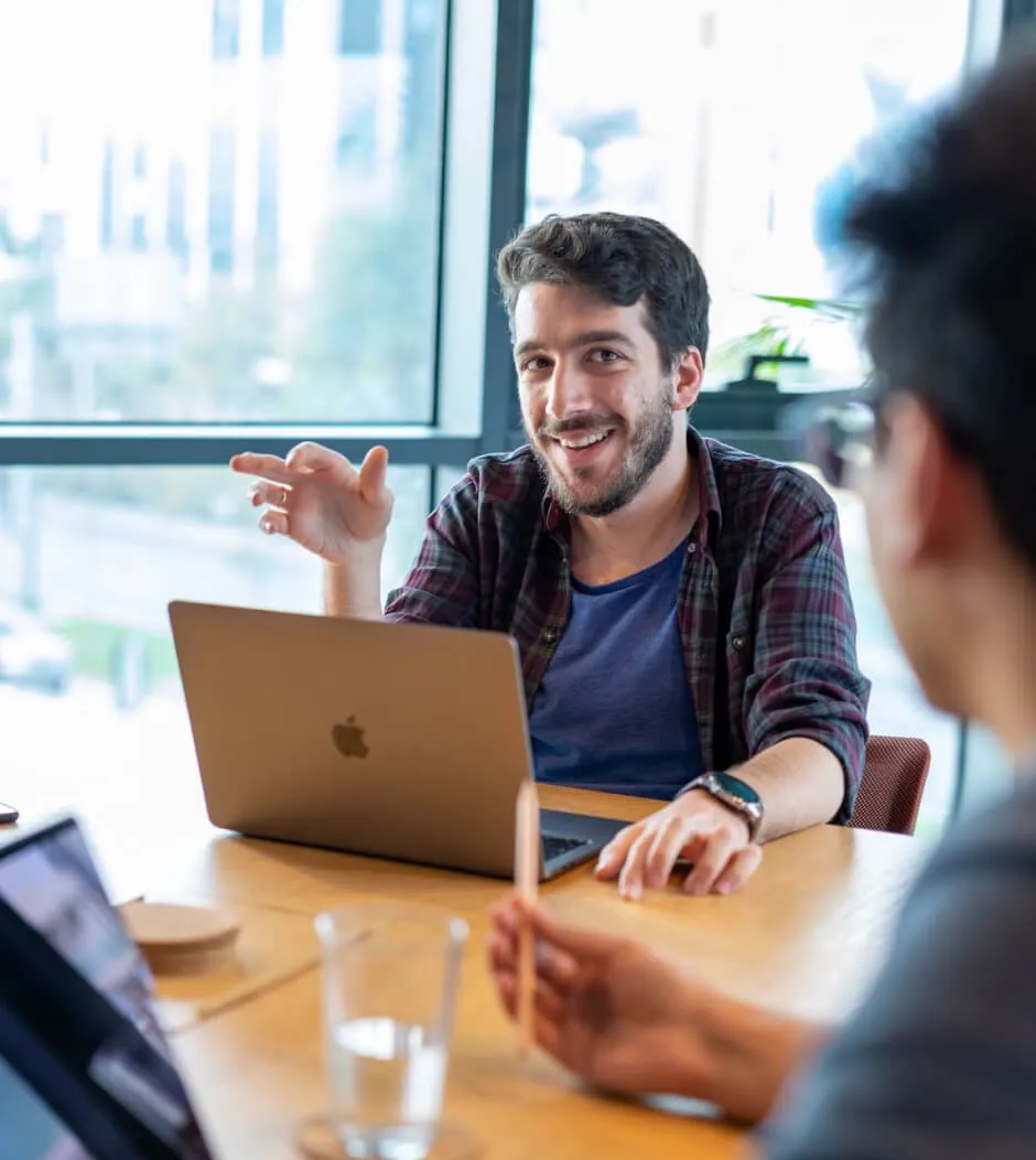 Man in a plaid shirt gesturing during a conversation, laptop open in front of him.