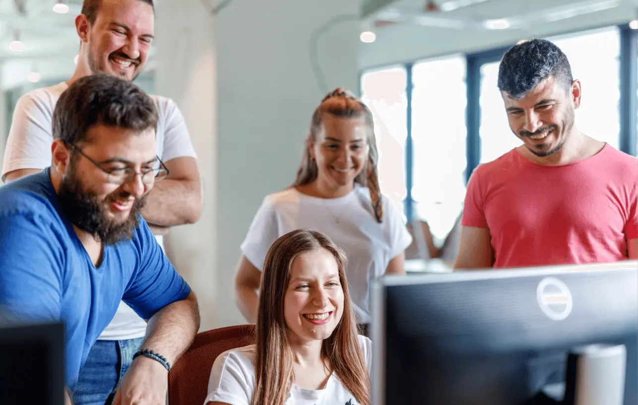 Team members gather around a computer, smiling and reacting to something on the screen.