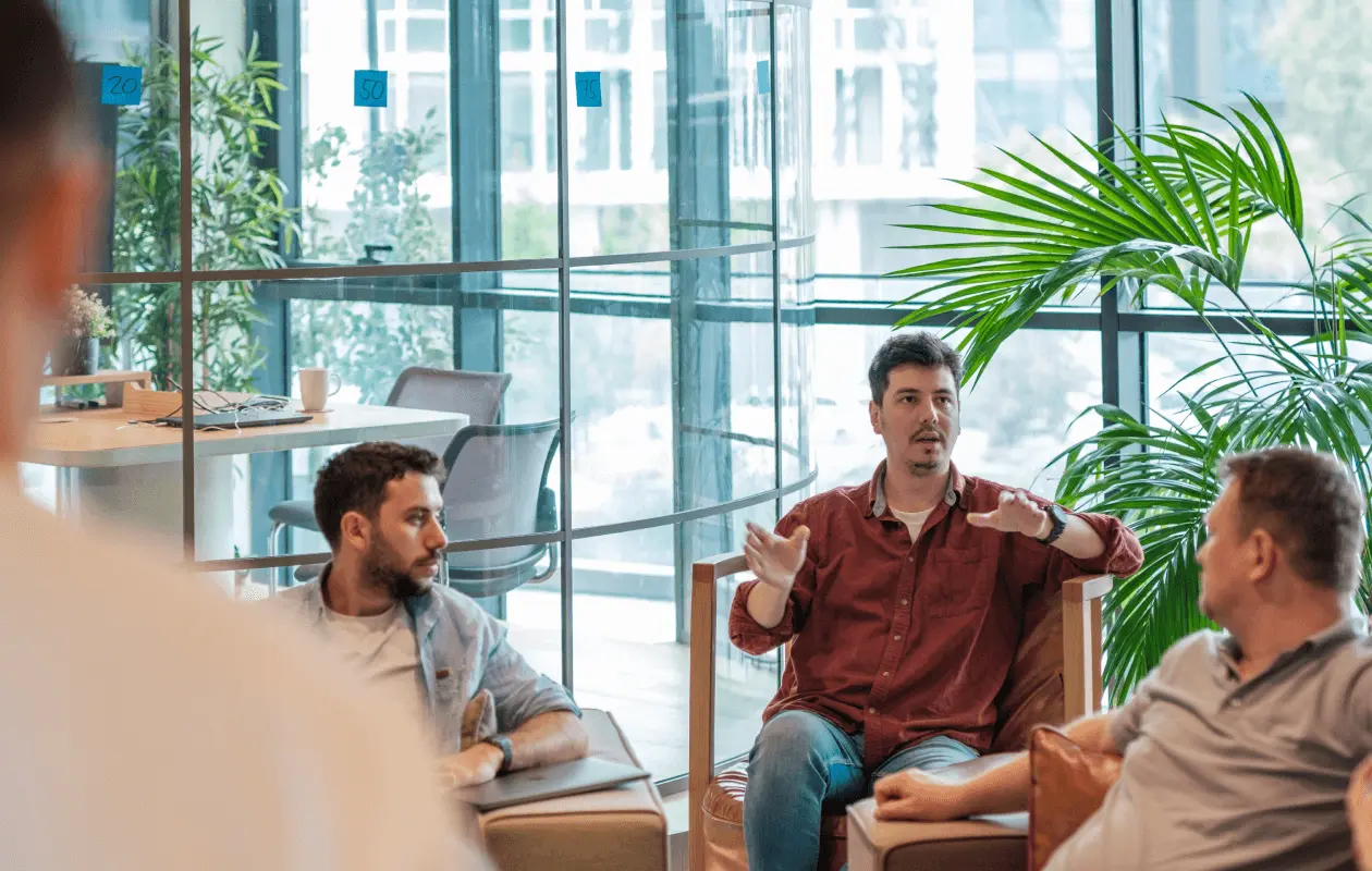 A man in a red shirt speaks while colleagues listen in a glass-walled office with plants.