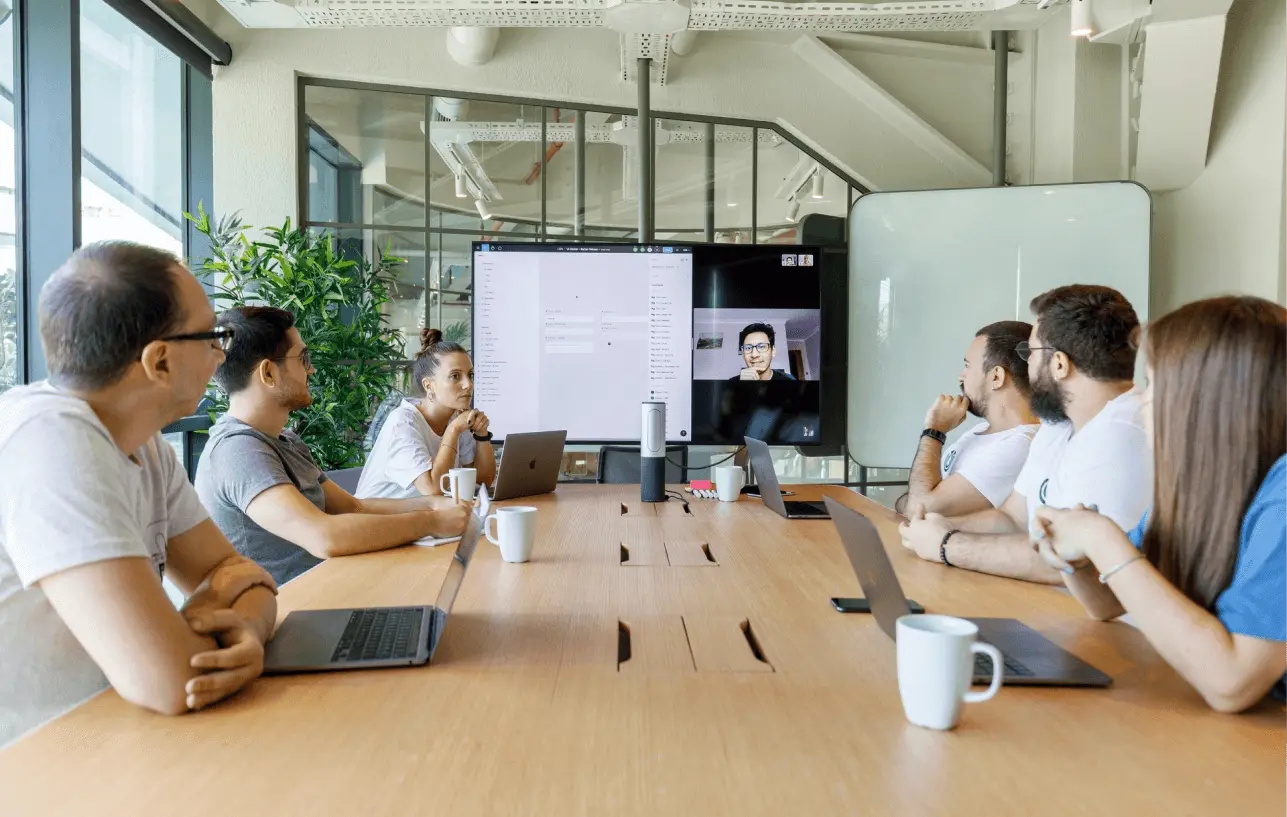 A team sits around a conference table, engaging in a video call with a remote colleague.