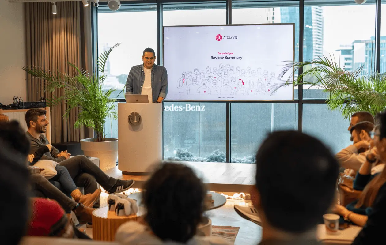 A team member presents a review summary to colleagues in a modern office, with an Atolye15 slide on the screen.