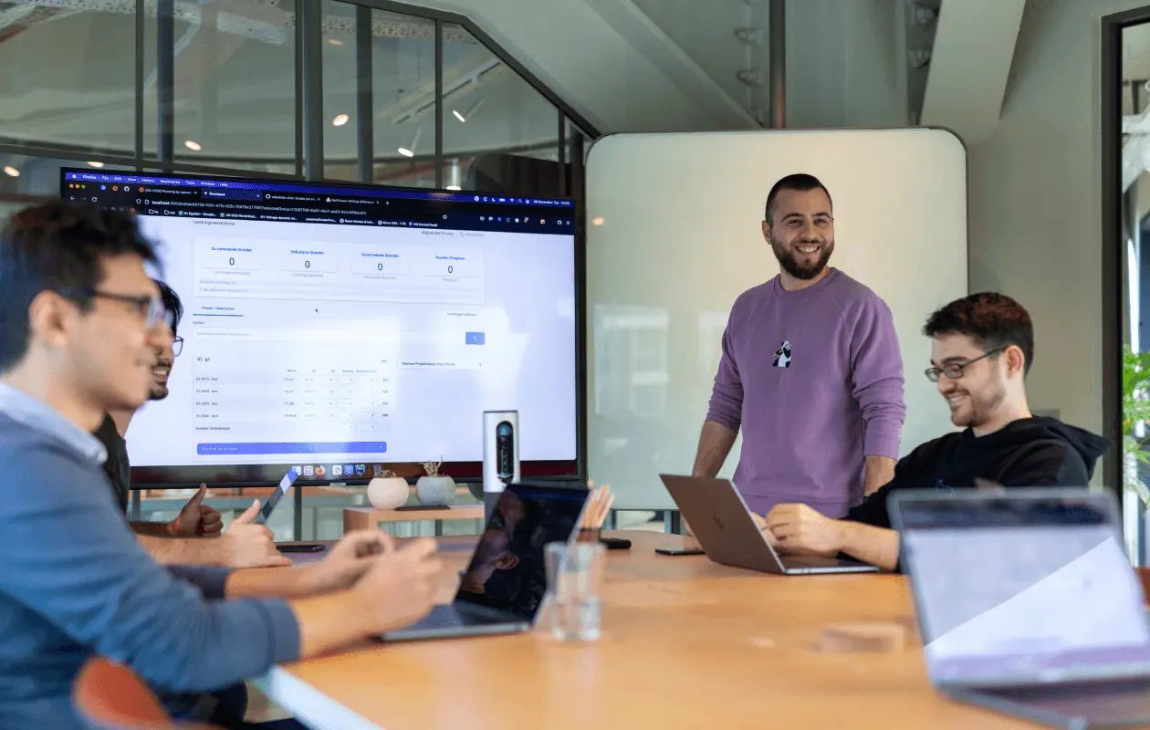 A smiling presenter leads a discussion in front of a screen displaying data, with teammates engaged.