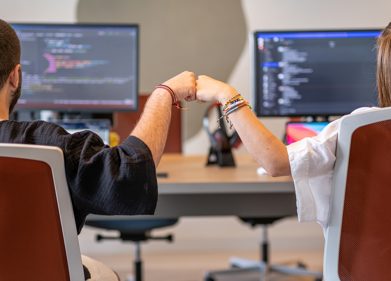 Two developers fist-bumping at their desks with code visible on monitors.