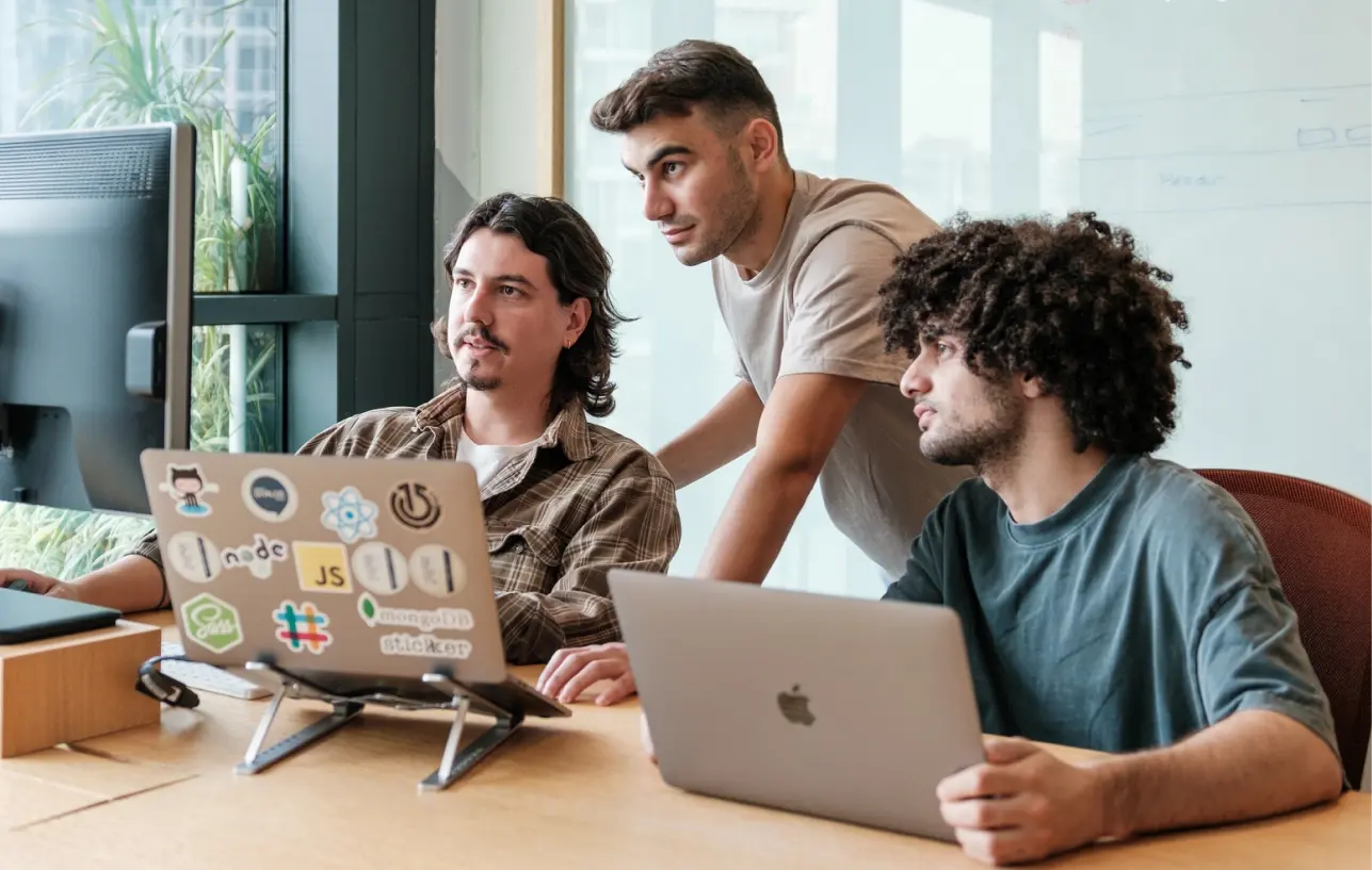 Three developers collaborate around their laptops in a bright office space, reviewing code on screen.