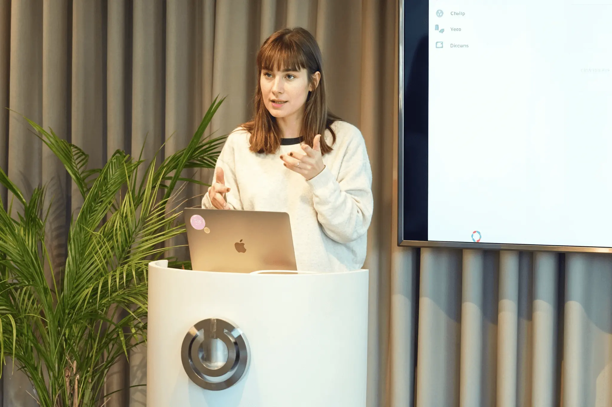 Woman giving a presentation behind a laptop at a podium, speaking to the audience.