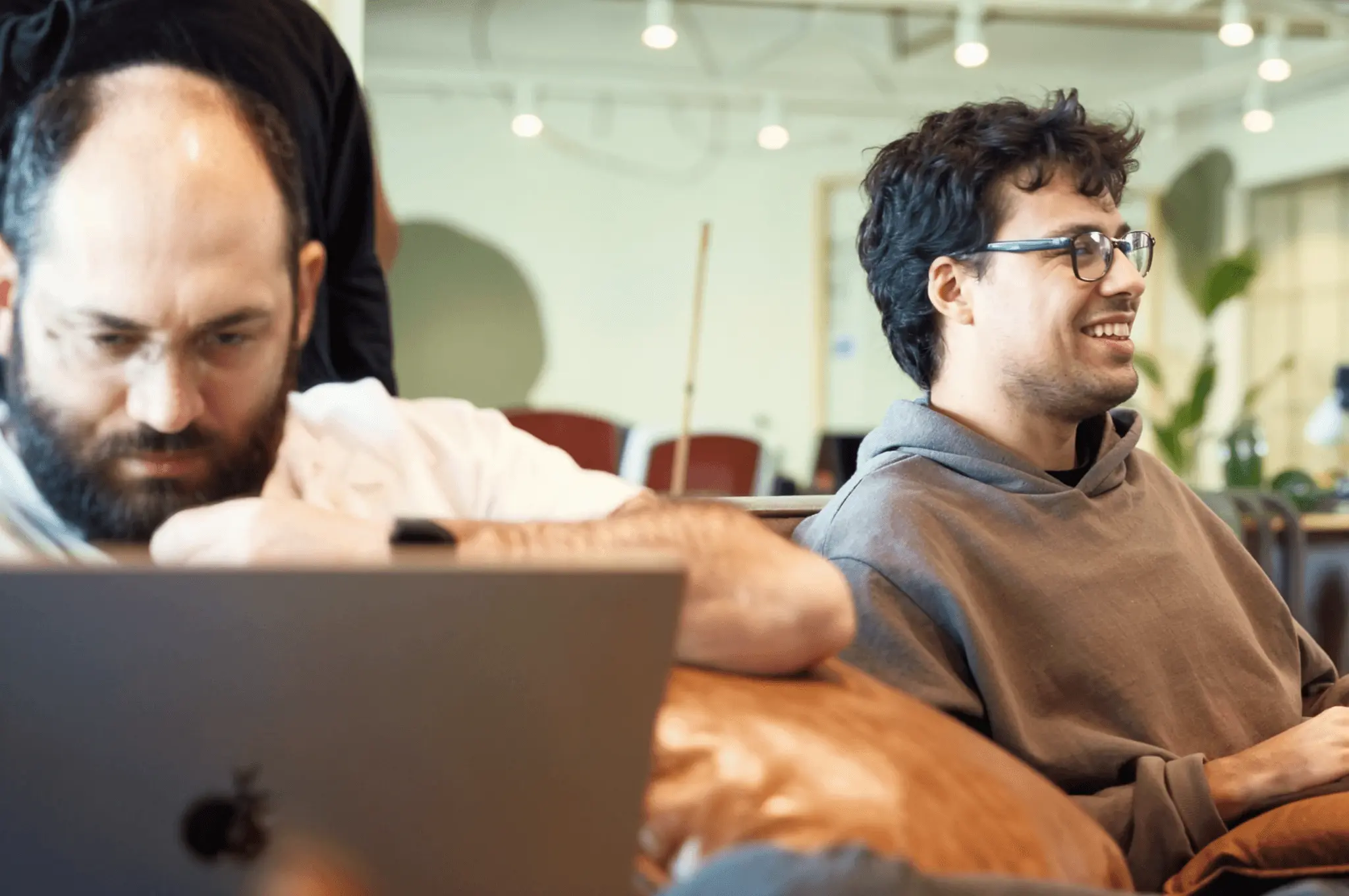 Developer smiling on a couch while another focuses on his laptop next to him.