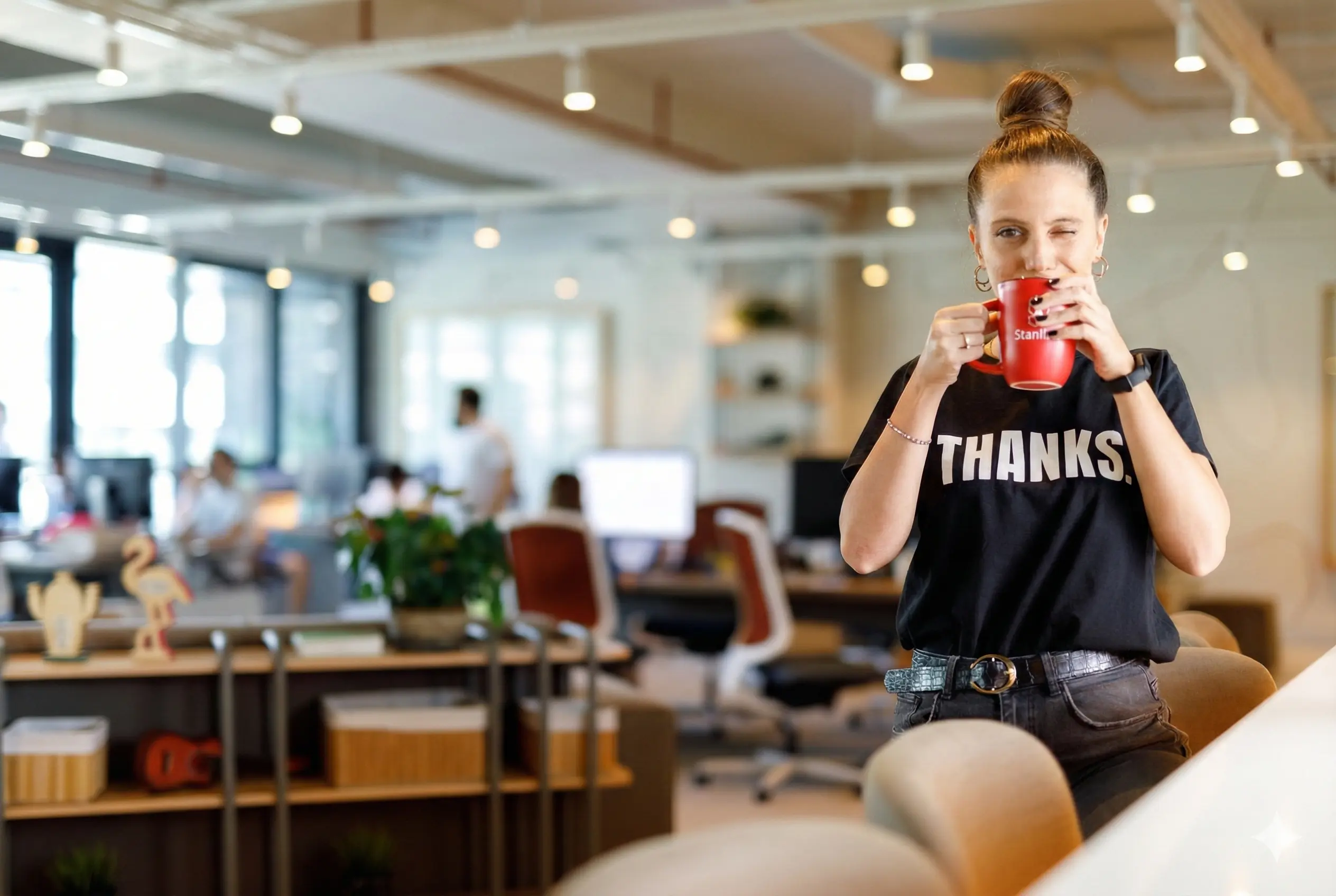 Woman in office holding red mug, winking, wearing “THANKS.” shirt